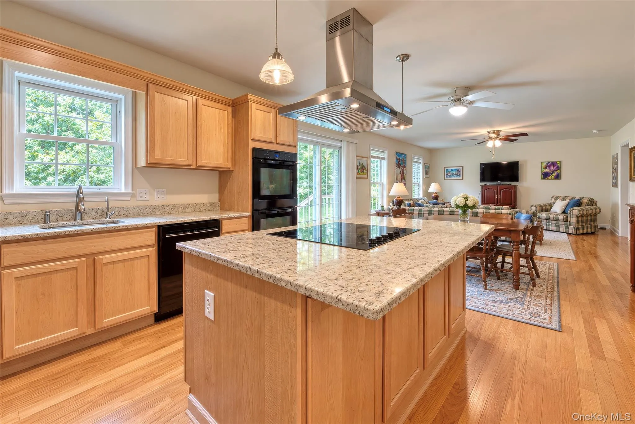 Open-concept kitchen featuring an island with a built-in cooktop, granite countertops, wood-finish flooring, light wood cabinetry, and a stainless steel range hood Open-concept kitchen featuring an island with a built-in cooktop, granite countertops, wood-finish flooring, light wood cabinetry, and a stainless steel range hood