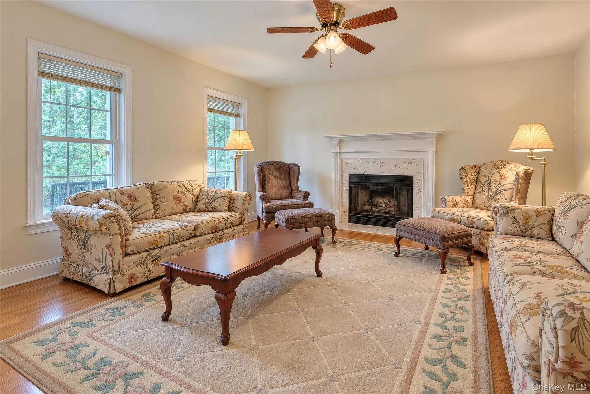Hardwood flooring, white window trim, fireplace with a white mantel, ceiling fan with integrated lighting, and light-colored wall paint Hardwood flooring, white window trim, fireplace with a white mantel, ceiling fan with integrated lighting, and light-colored wall paint