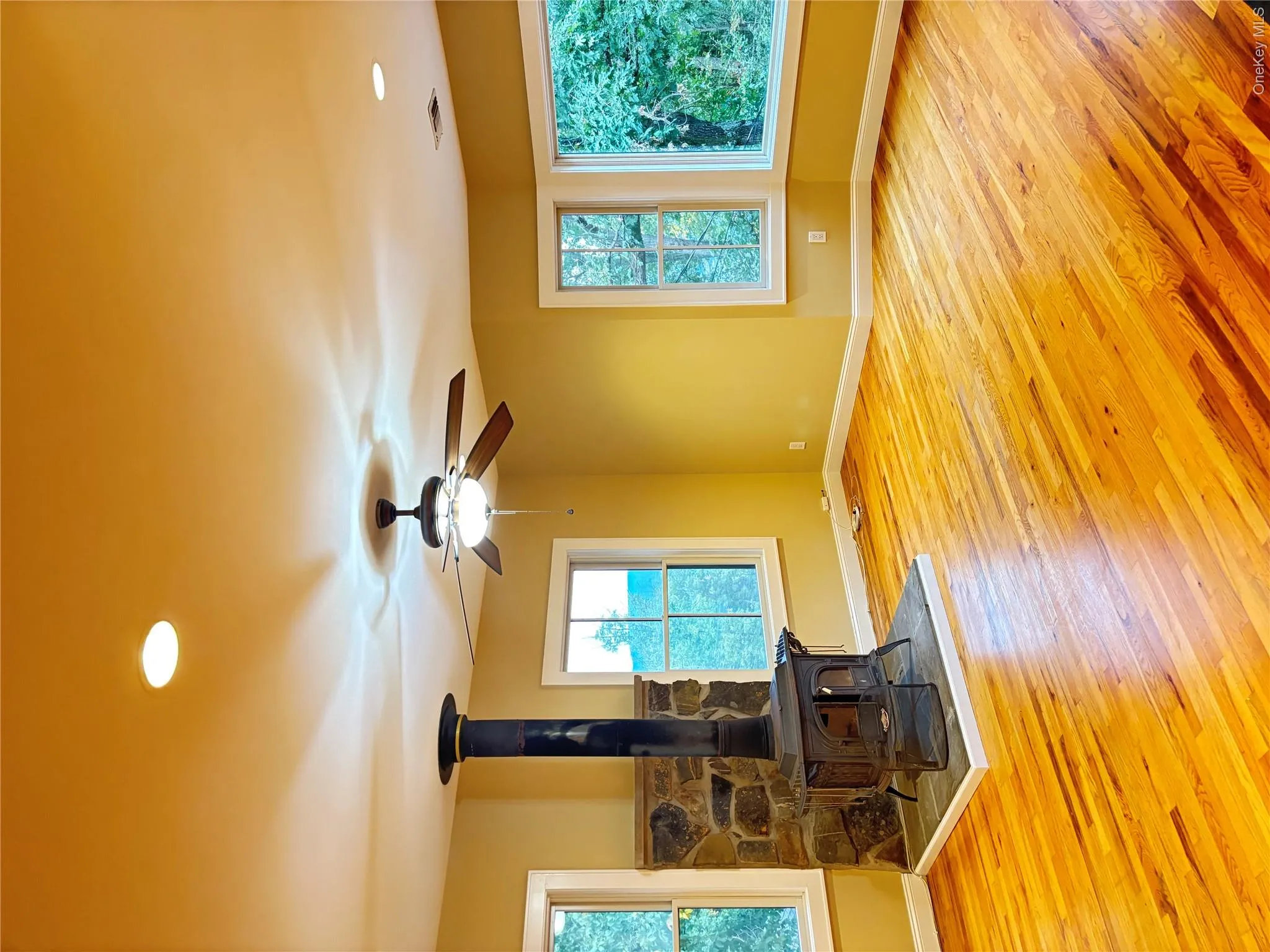 Thus spacious living room has hardwood flooring, wood-burning stove with stone surround, ceiling fan, recessed lighting, and multiple windows. Thus spacious living room has hardwood flooring, wood-burning stove with stone surround, ceiling fan, recessed lighting, and multiple windows.