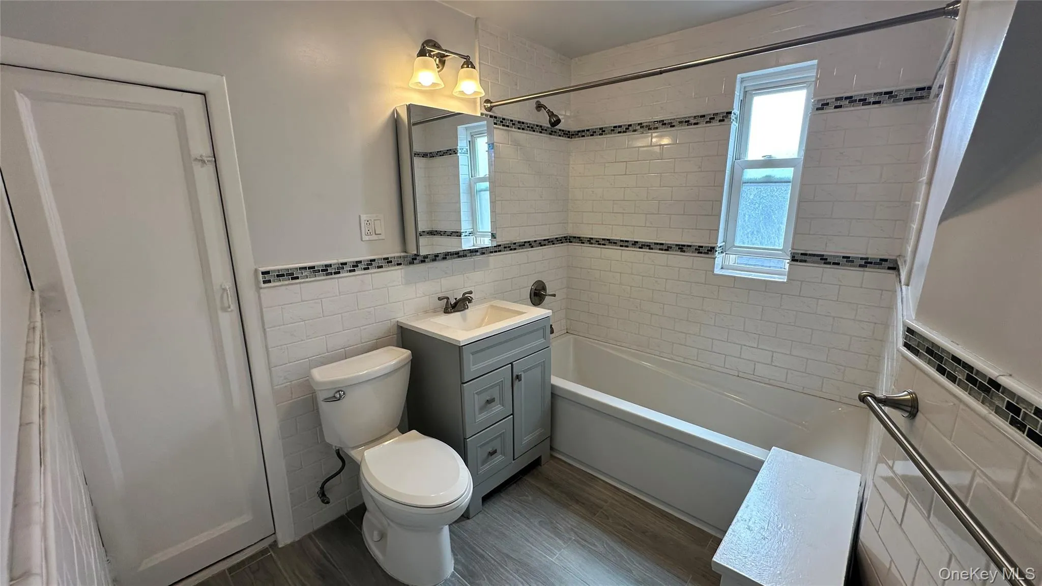 White subway tile with decorative mosaic accents, gray wood-finish flooring, a contemporary gray vanity with sink, and a mirrored medicine cabinet White subway tile with decorative mosaic accents, gray wood-finish flooring, a contemporary gray vanity with sink, and a mirrored medicine cabinet
