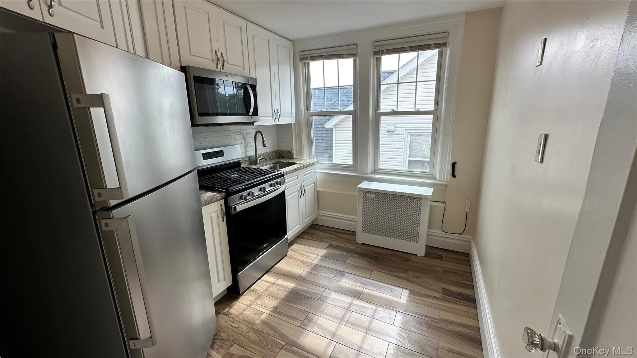 Kitchen featuring stainless steel appliances, white cabinetry, wood-finish flooring, subway tile backsplash, and dual windows Kitchen featuring stainless steel appliances, white cabinetry, wood-finish flooring, subway tile backsplash, and dual windows