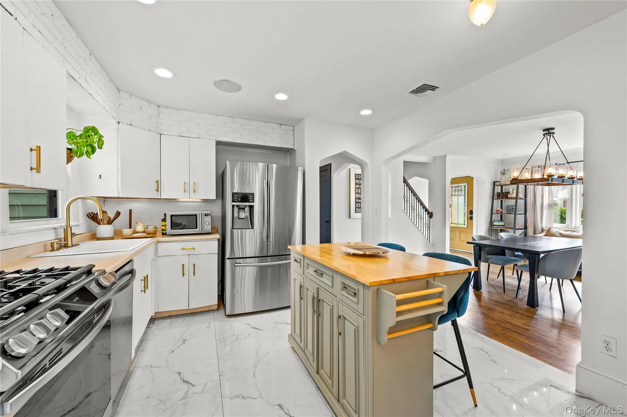 White cabinetry with gold-finish hardware, stainless steel appliances, a center island with a wood-finish countertop, marble-look floor tile, and an arched entryway to the dining area White cabinetry with gold-finish hardware, stainless steel appliances, a center island with a wood-finish countertop, marble-look floor tile, and an arched entryway to the dining area