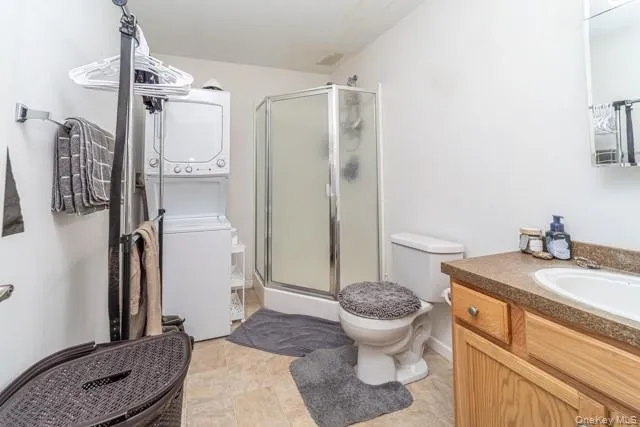 Compact bathroom featuring a shower stall with clear glass enclosure, a vanity with wood-finish cabinetry, a built-in sink, and a stacked washer and dryer unit Compact bathroom featuring a shower stall with clear glass enclosure, a vanity with wood-finish cabinetry, a built-in sink, and a stacked washer and dryer unit