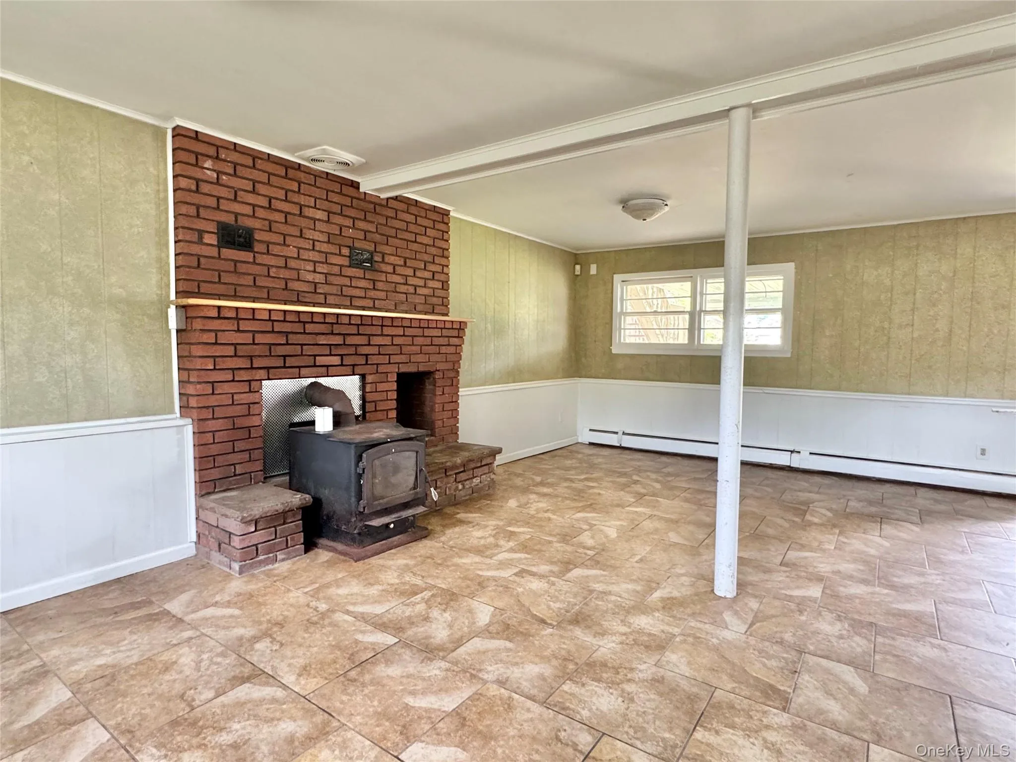 Unfurnished living room with a wood stove, wainscoting, beamed ceiling, ornamental molding, and a baseboard heating unit Unfurnished living room with a wood stove, wainscoting, beamed ceiling, ornamental molding, and a baseboard heating unit