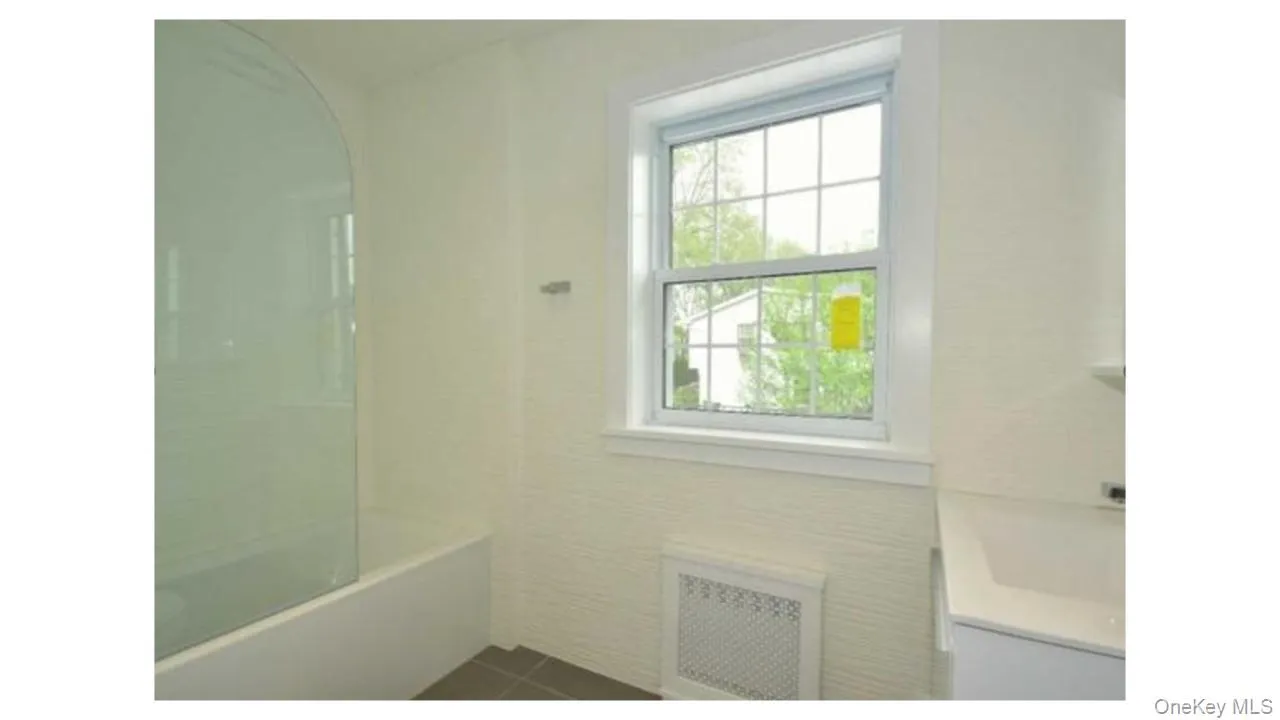 White bathtub with a curved glass shower partition, white window with grid panes, textured wall covering, and a white vanity with an integrated sink White bathtub with a curved glass shower partition, white window with grid panes, textured wall covering, and a white vanity with an integrated sink