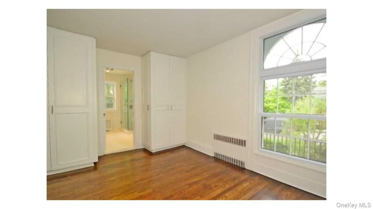 Wood-finish flooring with built-in cabinetry, an arched window, and an en-suite bathroom with a glass-enclosed shower Wood-finish flooring with built-in cabinetry, an arched window, and an en-suite bathroom with a glass-enclosed shower