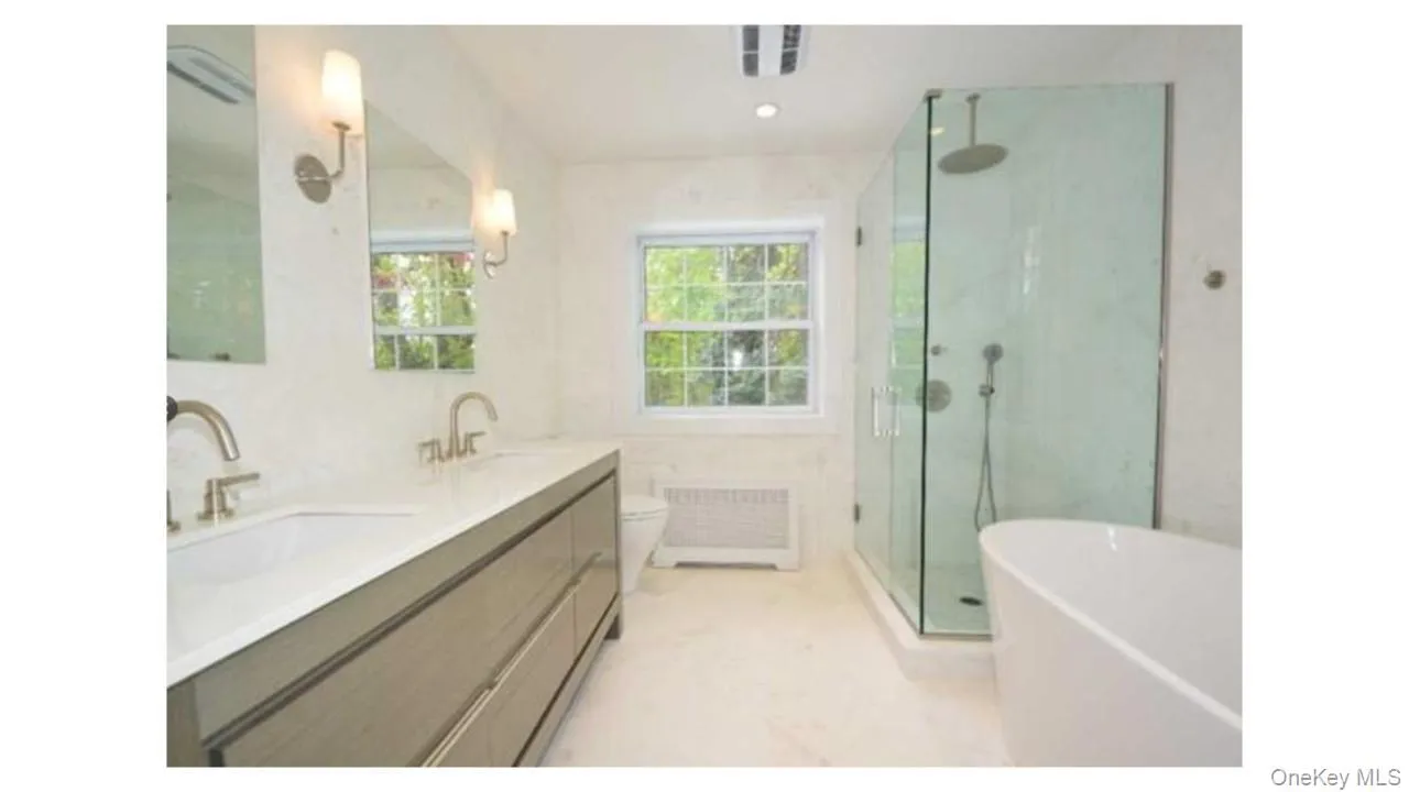 Dual vanity with integrated sinks, frameless glass shower enclosure, freestanding soaking tub, and natural light from a double-hung window Dual vanity with integrated sinks, frameless glass shower enclosure, freestanding soaking tub, and natural light from a double-hung window