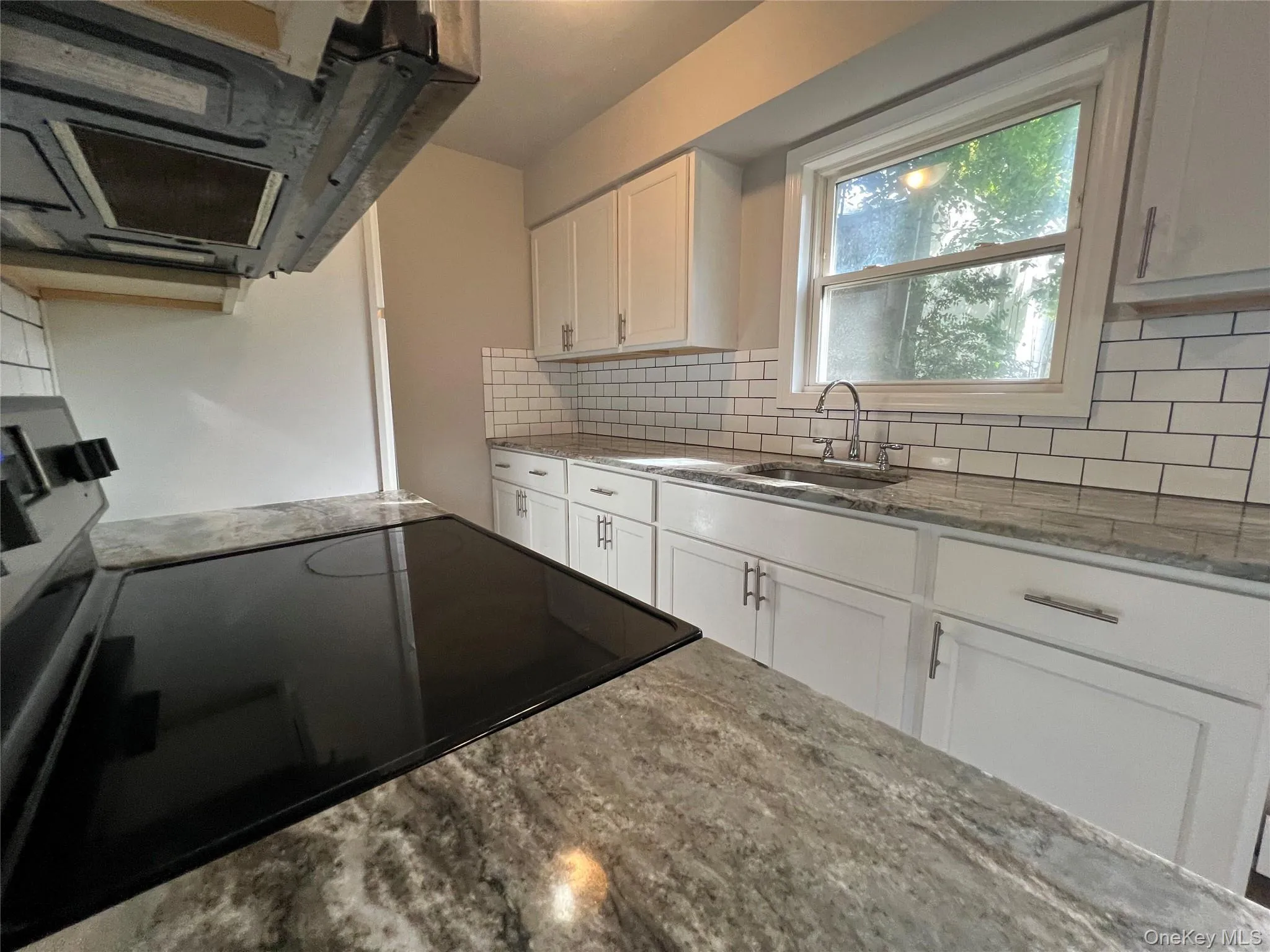 White shaker cabinetry, subway tile backsplash, integrated sink with chrome faucet, light-toned countertop, and a built-in cooktop White shaker cabinetry, subway tile backsplash, integrated sink with chrome faucet, light-toned countertop, and a built-in cooktop