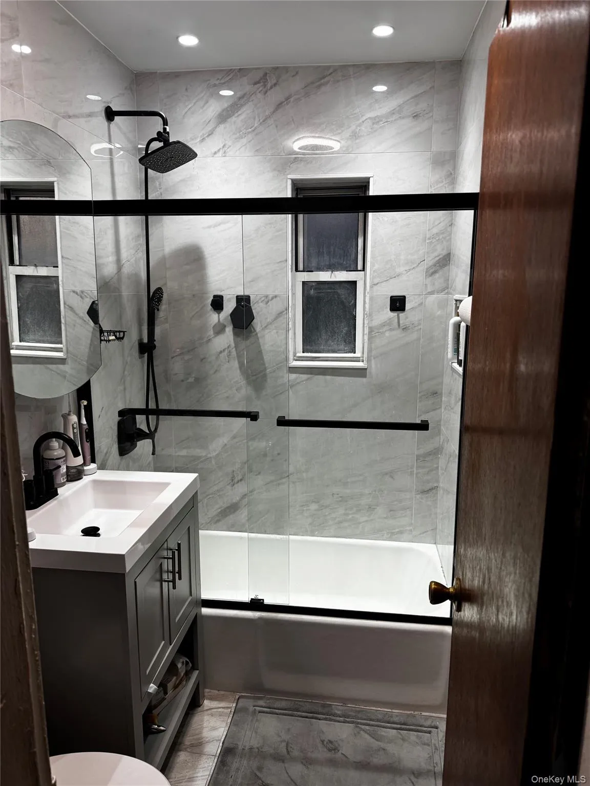 Bathroom featuring a white bathtub, glass shower enclosure with matte black hardware, gray vanity with integrated sink, and recessed lighting Bathroom featuring a white bathtub, glass shower enclosure with matte black hardware, gray vanity with integrated sink, and recessed lighting