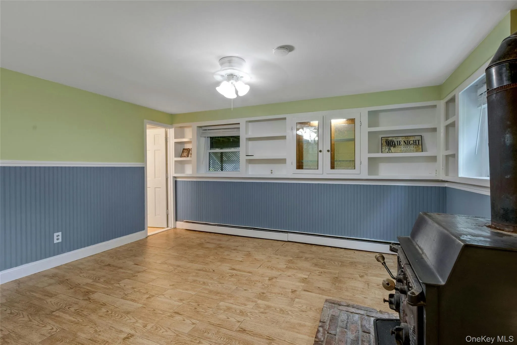 Wood-finish flooring with a ceiling fan, built-in shelving, windows, and a wood-burning stove with a brick hearth Wood-finish flooring with a ceiling fan, built-in shelving, windows, and a wood-burning stove with a brick hearth