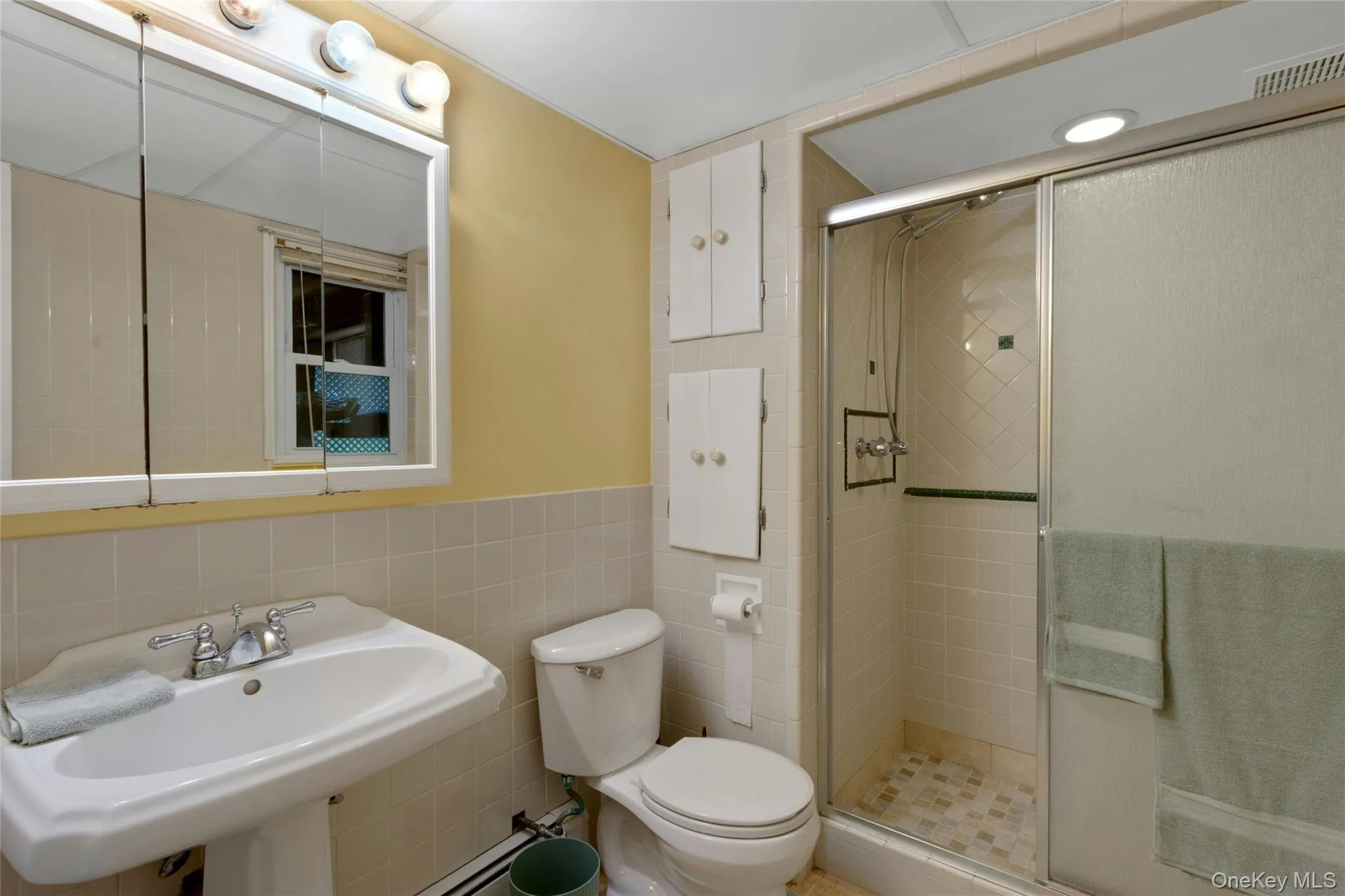 Pedestal sink, wall-mounted medicine cabinet with integrated lighting, tiled shower with glass enclosure, recessed shower light, and built-in wall cabinetry Pedestal sink, wall-mounted medicine cabinet with integrated lighting, tiled shower with glass enclosure, recessed shower light, and built-in wall cabinetry