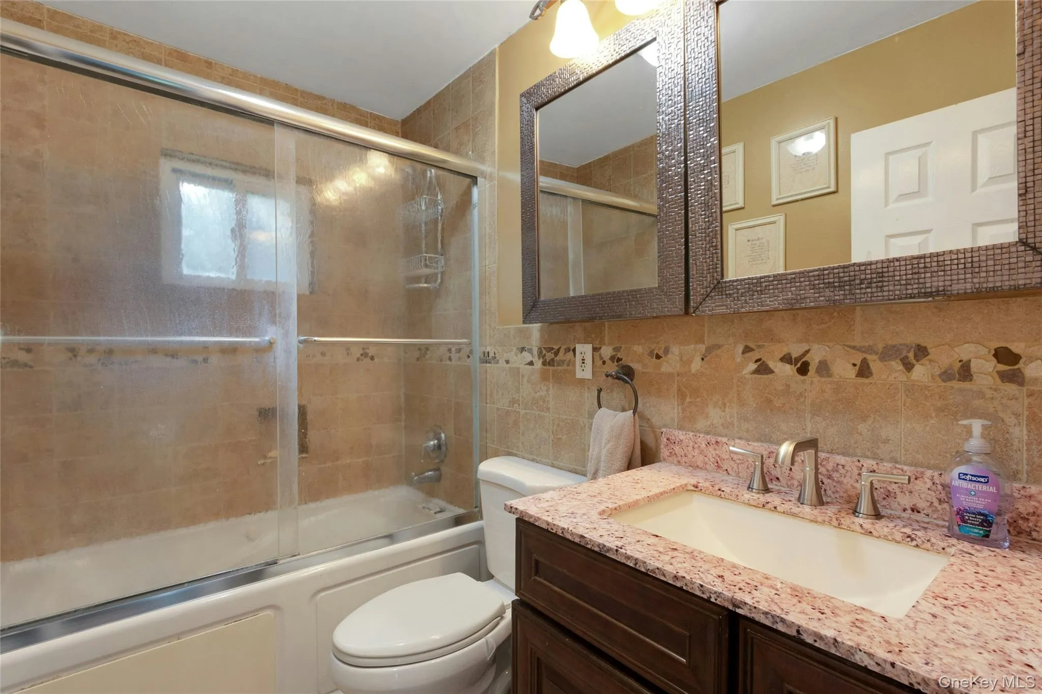Bathroom featuring a built-in tub with glass sliding doors, a vanity with a light-colored countertop, an integrated sink, a framed mirror, and tiled walls with decorative accents Bathroom featuring a built-in tub with glass sliding doors, a vanity with a light-colored countertop, an integrated sink, a framed mirror, and tiled walls with decorative accents