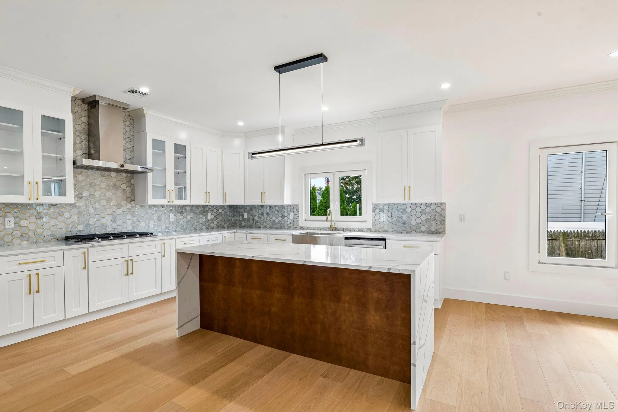 White shaker cabinetry with gold-tone hardware, hexagonal tile backsplash, stainless steel range hood, integrated kitchen island with contrasting wood-finish base, and light wood-finish flooring White shaker cabinetry with gold-tone hardware, hexagonal tile backsplash, stainless steel range hood, integrated kitchen island with contrasting wood-finish base, and light wood-finish flooring