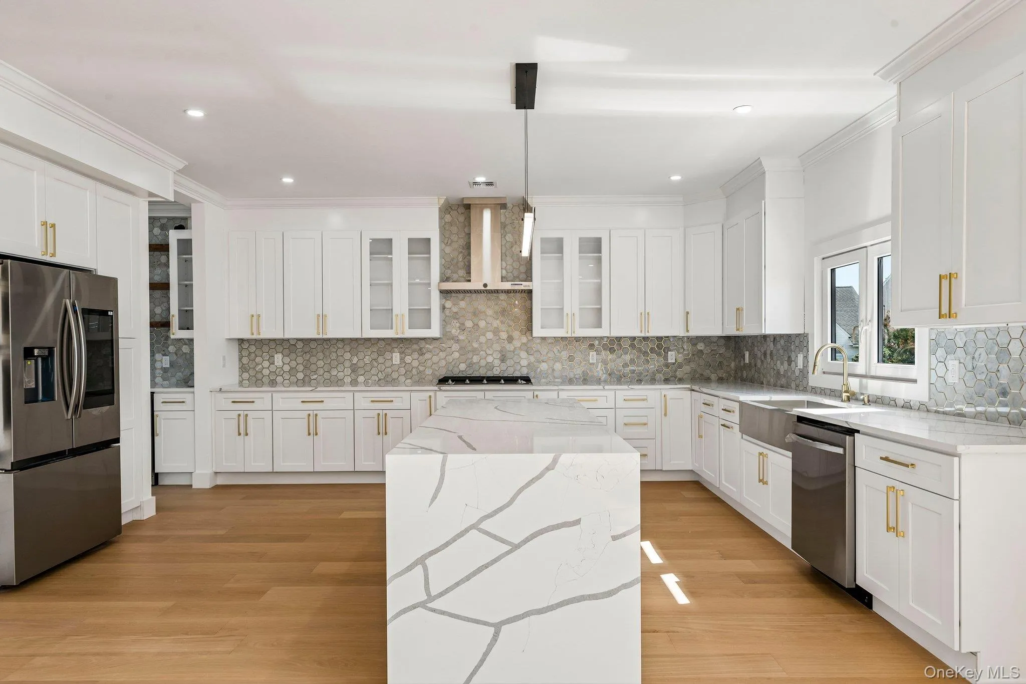 Gourmet kitchen featuring white shaker cabinetry, stainless steel appliances, a waterfall island, hexagon tile backsplash, and wood-finish flooring Gourmet kitchen featuring white shaker cabinetry, stainless steel appliances, a waterfall island, hexagon tile backsplash, and wood-finish flooring
