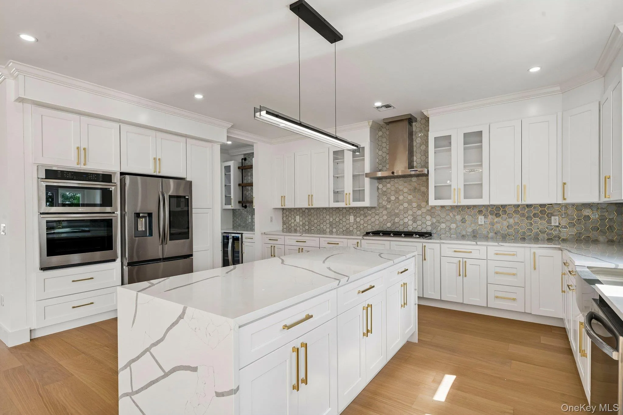 Gourmet kitchen featuring a waterfall island, stainless steel appliances, honeycomb tile backsplash, white cabinetry with gold-tone hardware, and wood-finish flooring Gourmet kitchen featuring a waterfall island, stainless steel appliances, honeycomb tile backsplash, white cabinetry with gold-tone hardware, and wood-finish flooring