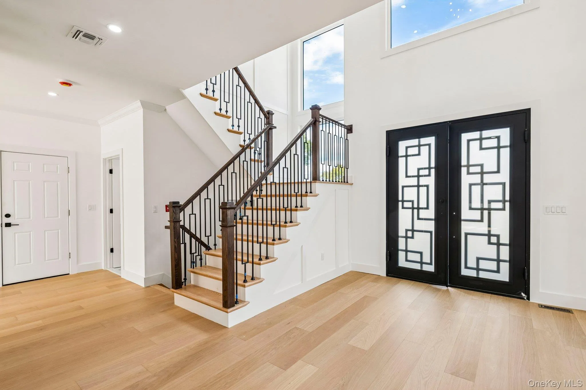 Two-story entryway with light wood-finish flooring, a contemporary black metal and wood staircase, and double entry doors with geometric glass inserts Two-story entryway with light wood-finish flooring, a contemporary black metal and wood staircase, and double entry doors with geometric glass inserts