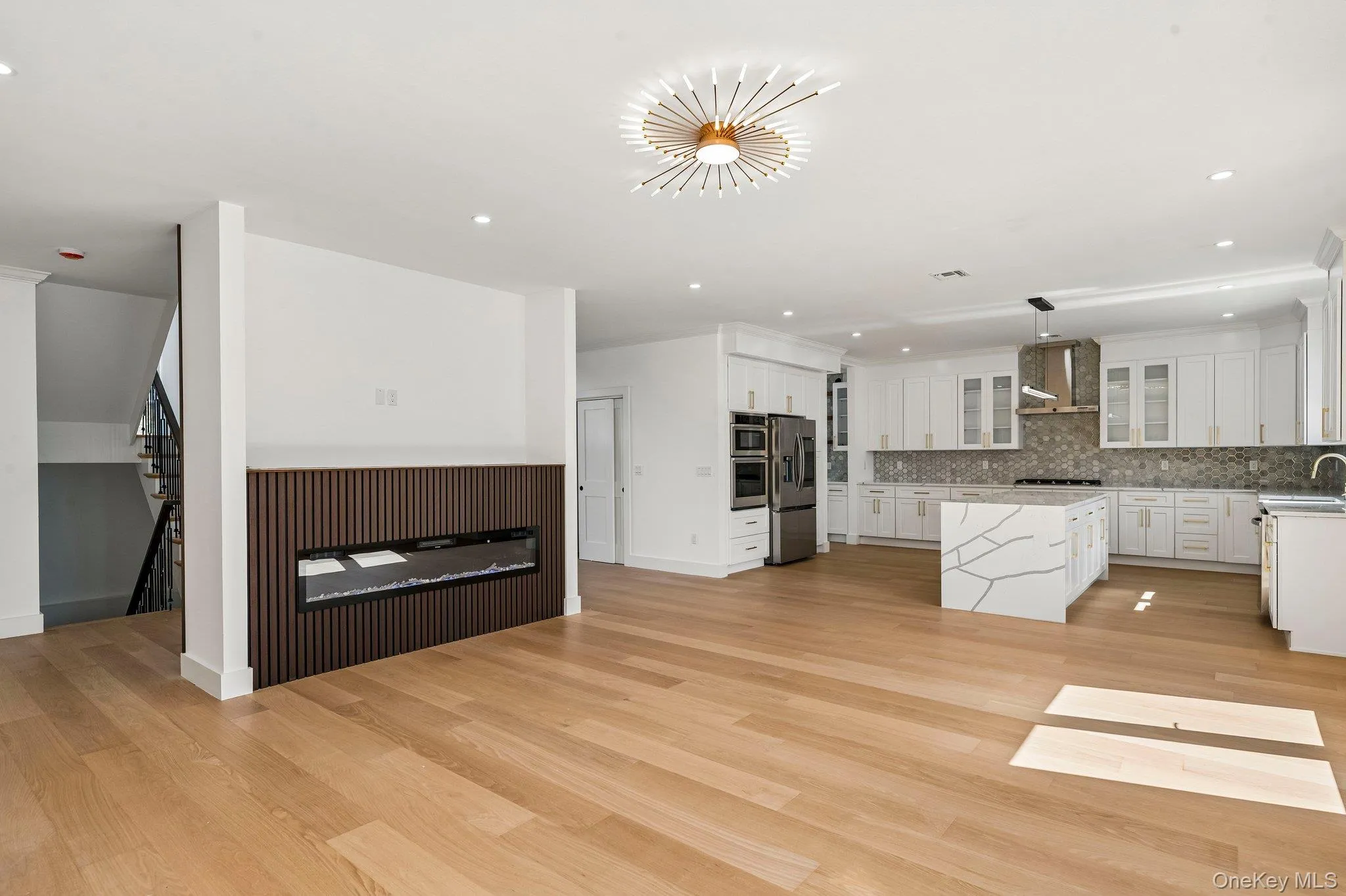 Light wood-finish flooring, built-in electric fireplace with slatted paneling, white shaker cabinetry, stainless steel appliances, and a kitchen island with a waterfall-edge countertop Light wood-finish flooring, built-in electric fireplace with slatted paneling, white shaker cabinetry, stainless steel appliances, and a kitchen island with a waterfall-edge countertop