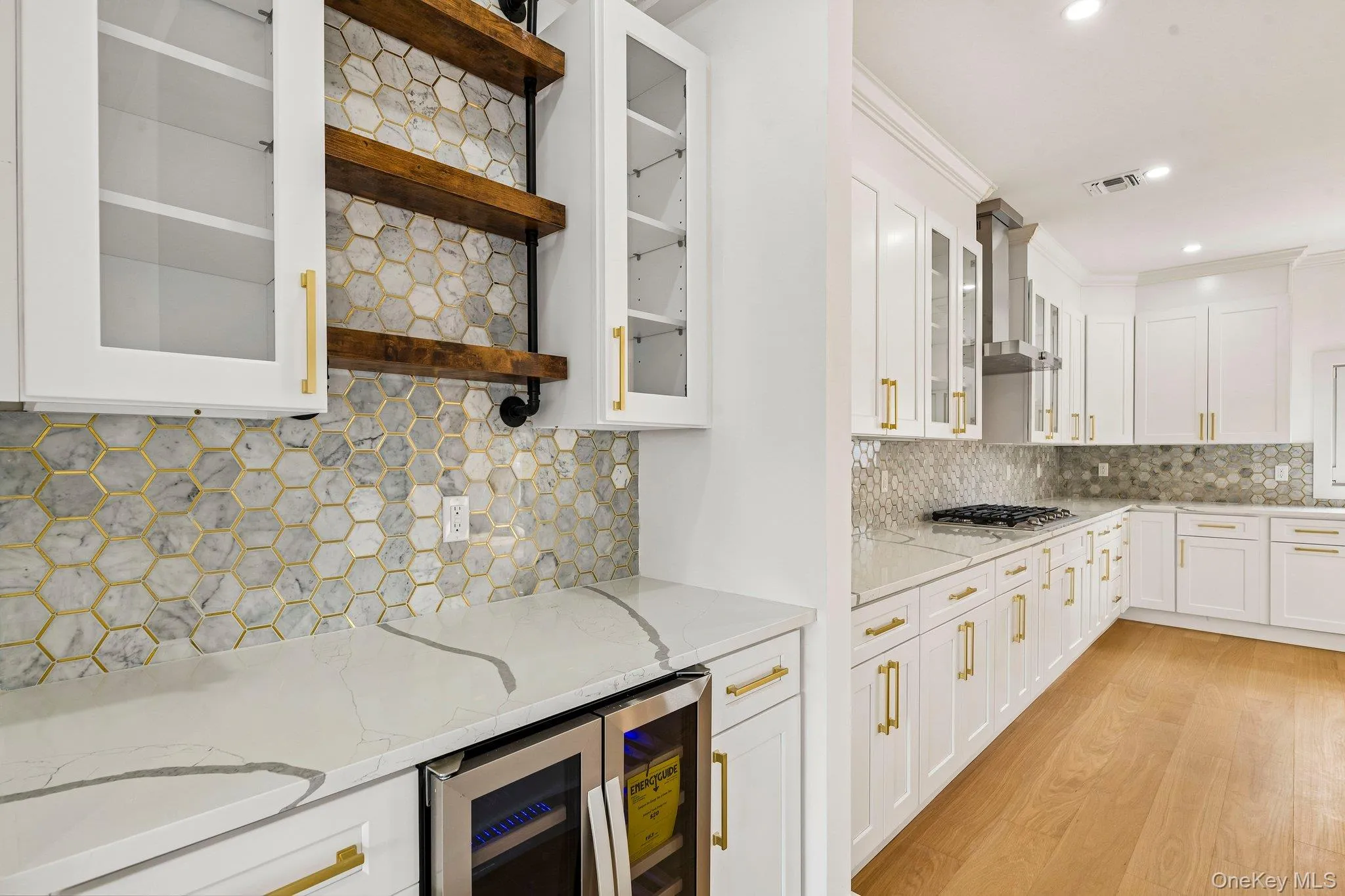 White cabinetry with gold-tone hardware, hexagon tile backsplash, quartz countertops, built-in beverage refrigerator, and wood-finish flooring White cabinetry with gold-tone hardware, hexagon tile backsplash, quartz countertops, built-in beverage refrigerator, and wood-finish flooring