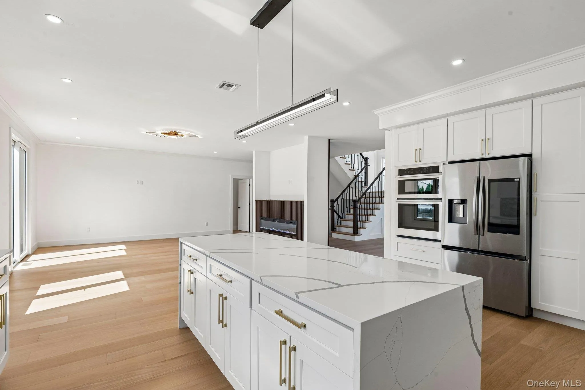 Kitchen island with waterfall countertop, stainless steel appliances, white cabinetry with gold-tone hardware, light wood-finish flooring, and recessed lighting Kitchen island with waterfall countertop, stainless steel appliances, white cabinetry with gold-tone hardware, light wood-finish flooring, and recessed lighting