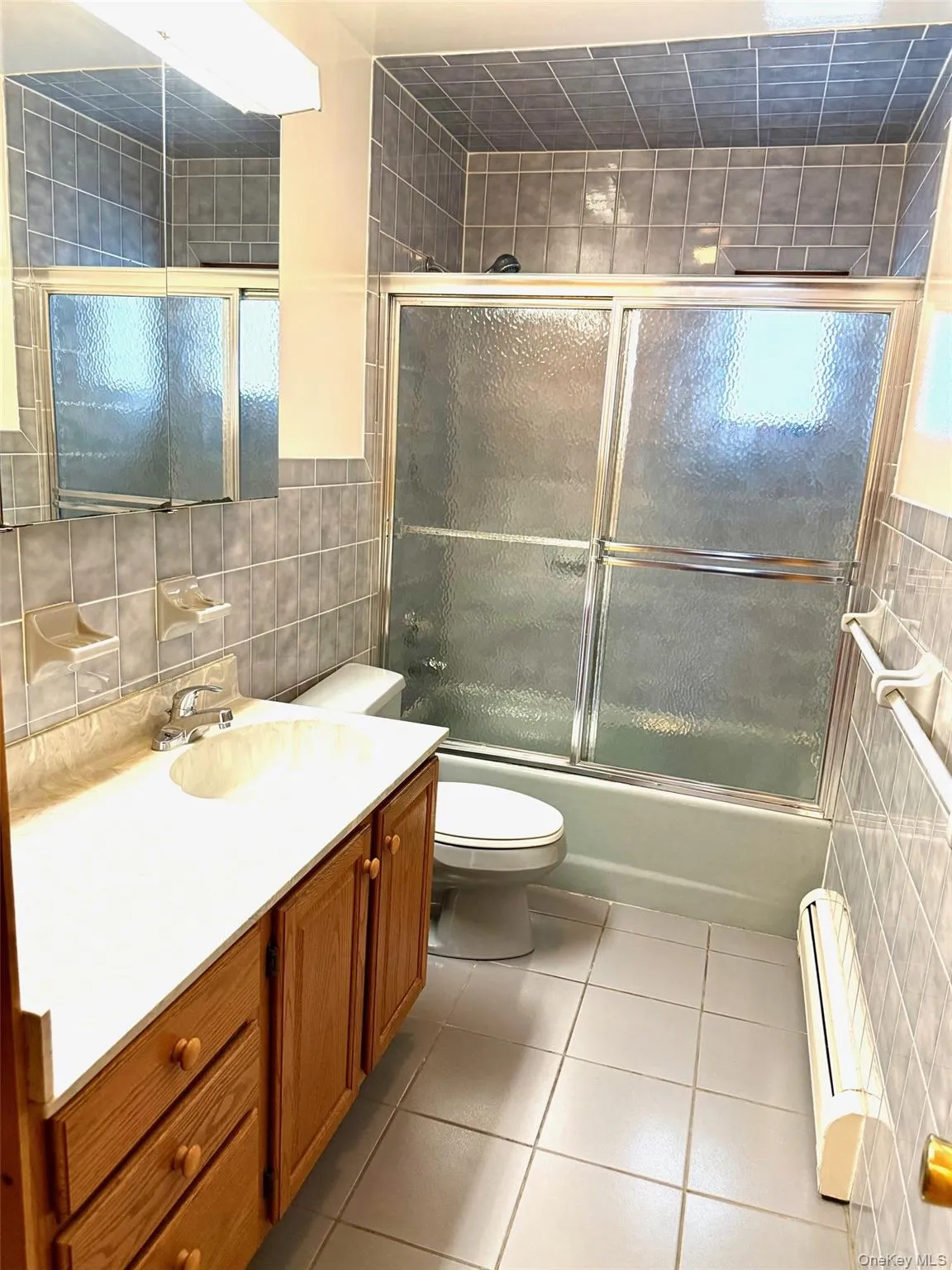 Hall Bathroom with tile walls, shower / bath combination with glass door, vanity, light tile patterned floors, and a baseboard radiator Hall Bathroom with tile walls, shower / bath combination with glass door, vanity, light tile patterned floors, and a baseboard radiator