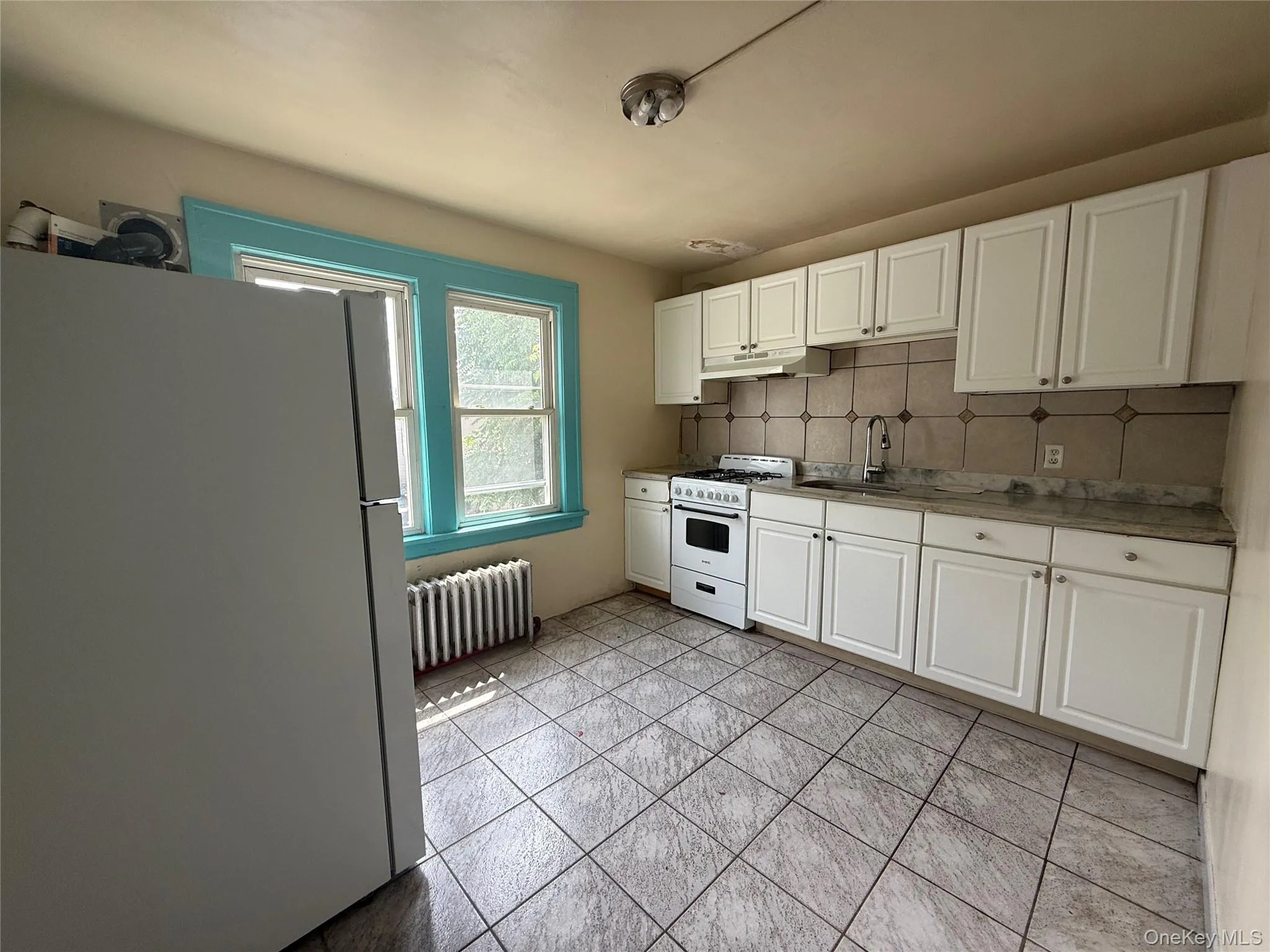 Kitchen featuring white appliances, white cabinetry, decorative backsplash, radiator, and under cabinet range hood Kitchen featuring white appliances, white cabinetry, decorative backsplash, radiator, and under cabinet range hood