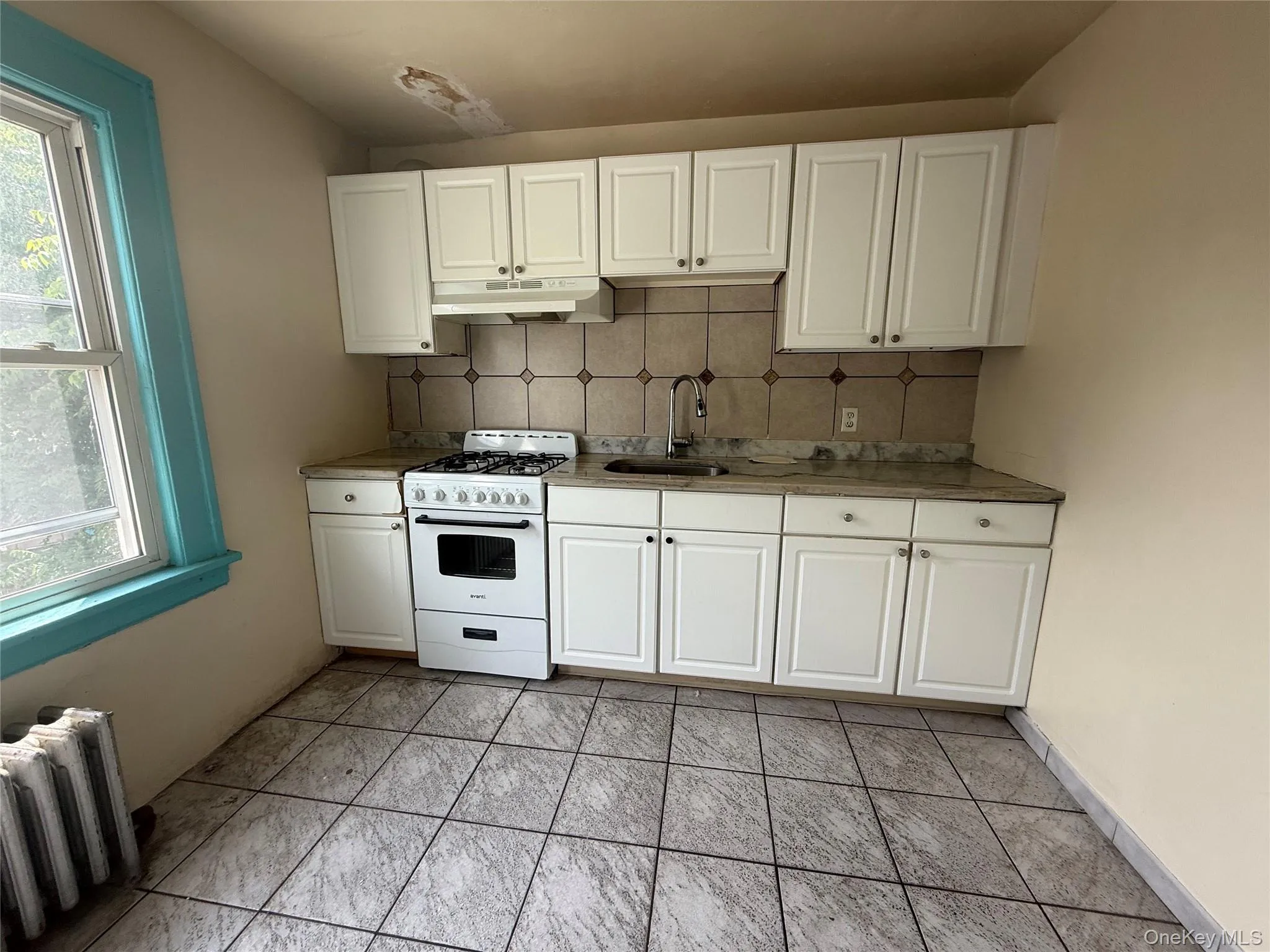 Kitchen with decorative backsplash, radiator heating unit, white cabinets, white range with gas cooktop, and under cabinet range hood Kitchen with decorative backsplash, radiator heating unit, white cabinets, white range with gas cooktop, and under cabinet range hood