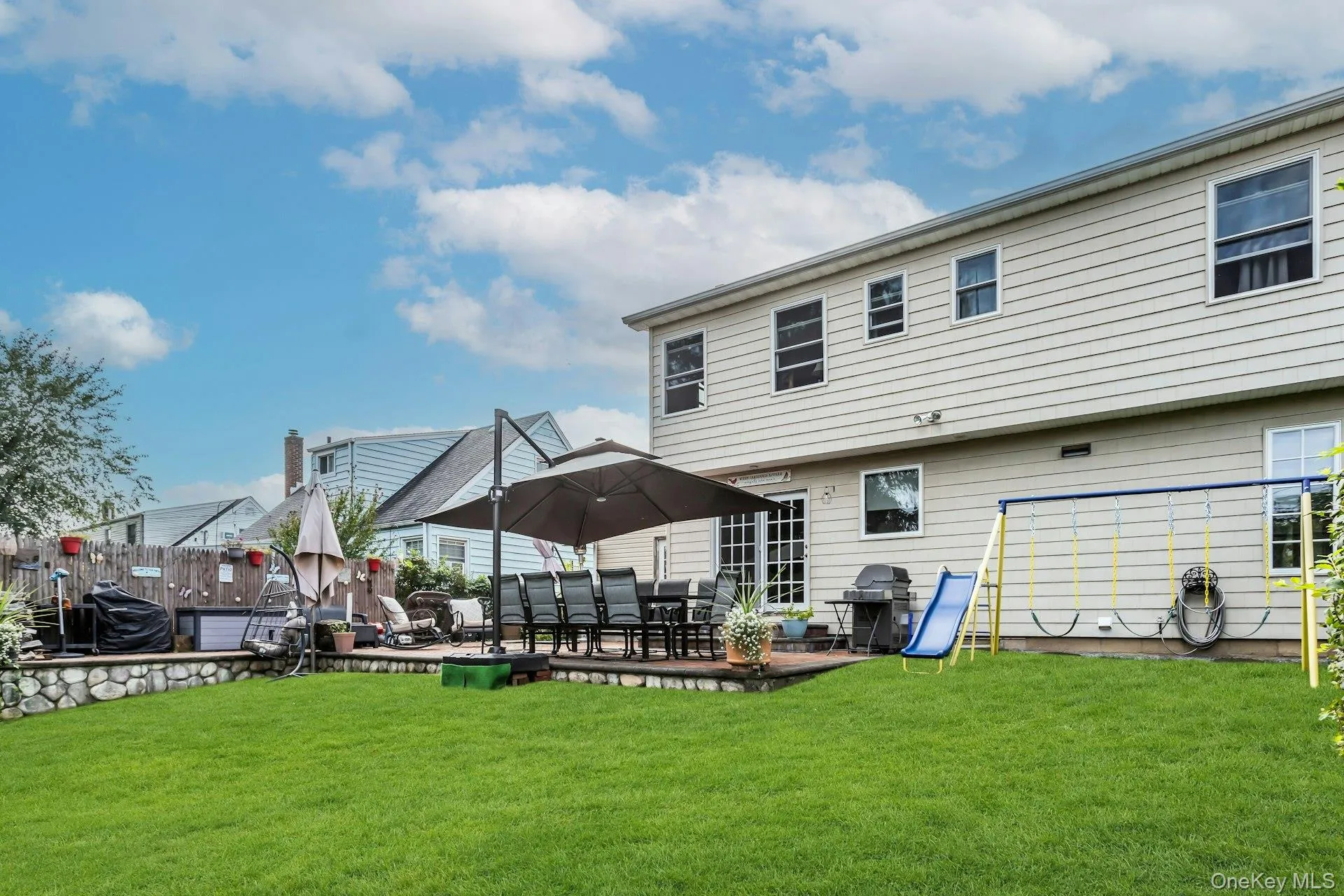 Expansive backyard featuring a manicured lawn, a raised stone-faced patio, and a two-story exterior with multiple windows Expansive backyard featuring a manicured lawn, a raised stone-faced patio, and a two-story exterior with multiple windows