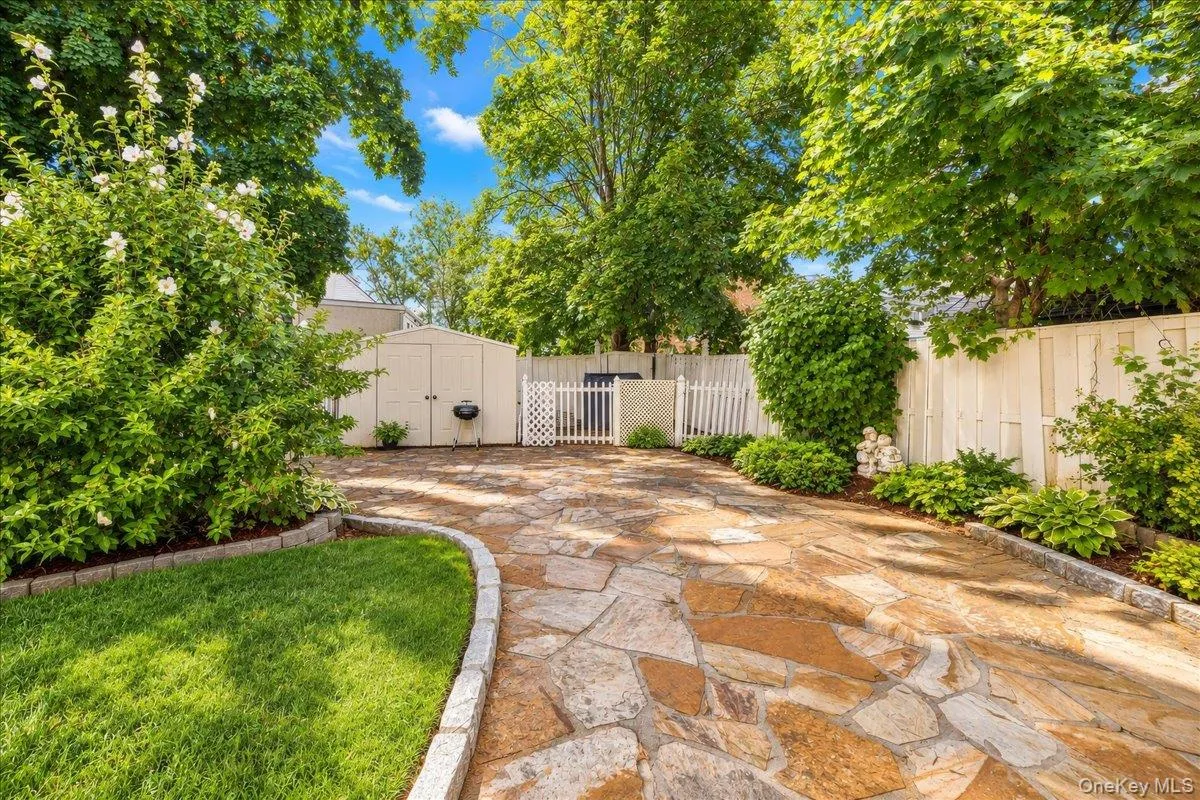 Expansive flagstone patio with a white picket fence, a storage shed, and mature landscaping Expansive flagstone patio with a white picket fence, a storage shed, and mature landscaping