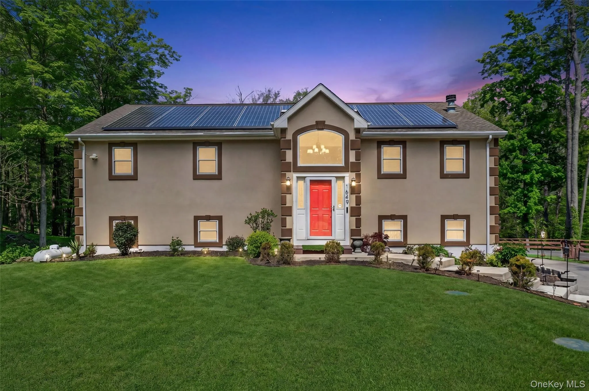 Stucco exterior with stone-look corner accents, solar panel array, arched entry window, red front door, and established landscaping Stucco exterior with stone-look corner accents, solar panel array, arched entry window, red front door, and established landscaping