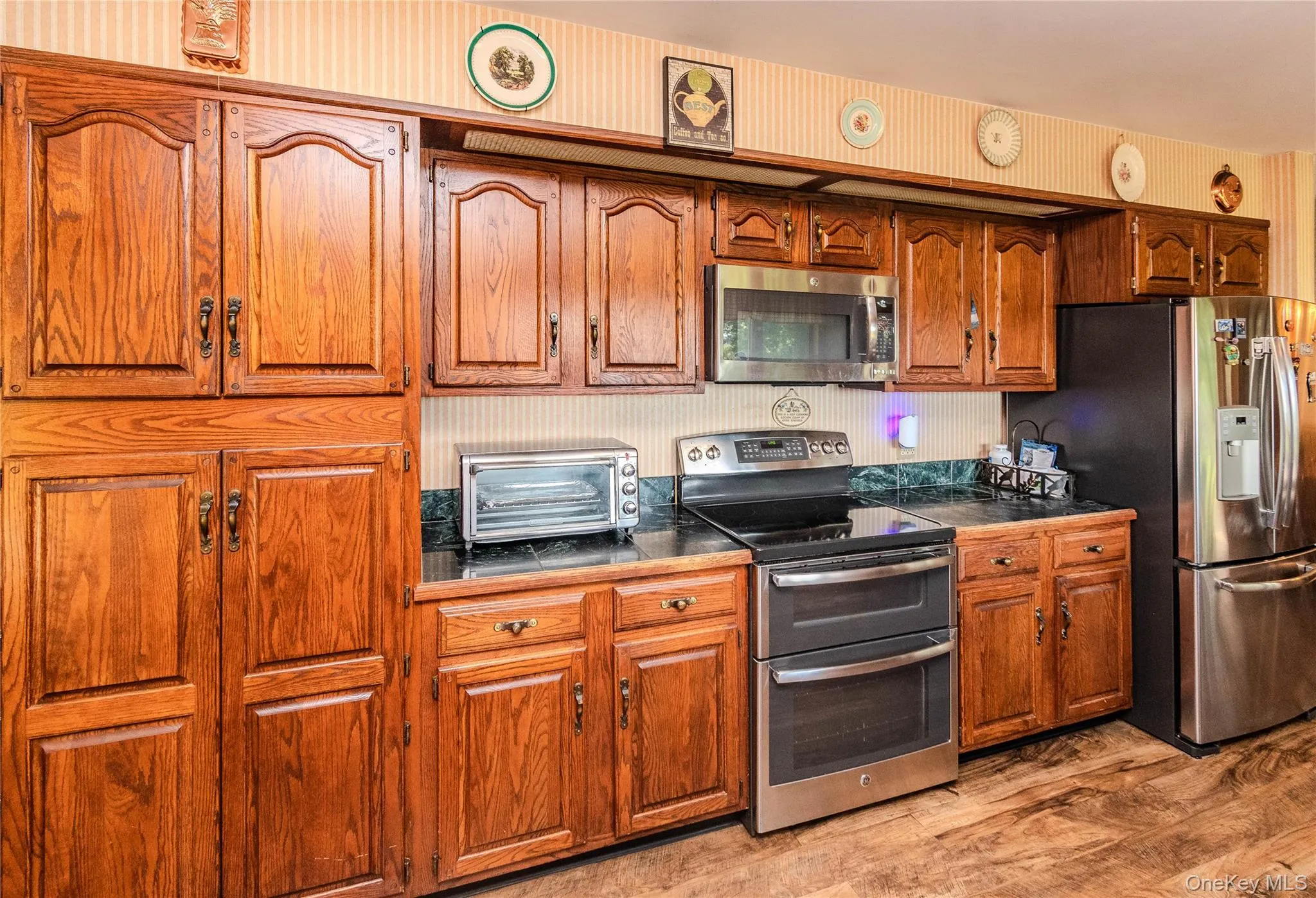 Wood-finish cabinetry with dark hardware, dark countertops, built-in stainless steel microwave, and wood-finish flooring Wood-finish cabinetry with dark hardware, dark countertops, built-in stainless steel microwave, and wood-finish flooring