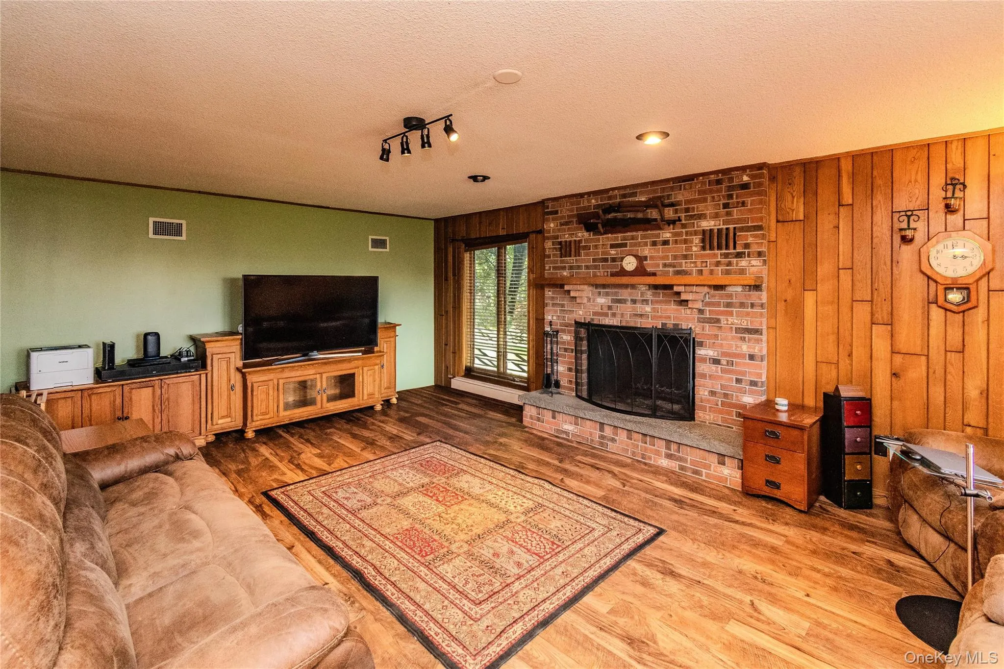 Wood-finish flooring throughout, featuring a brick fireplace with a stone hearth, wood paneling, and a large window Wood-finish flooring throughout, featuring a brick fireplace with a stone hearth, wood paneling, and a large window