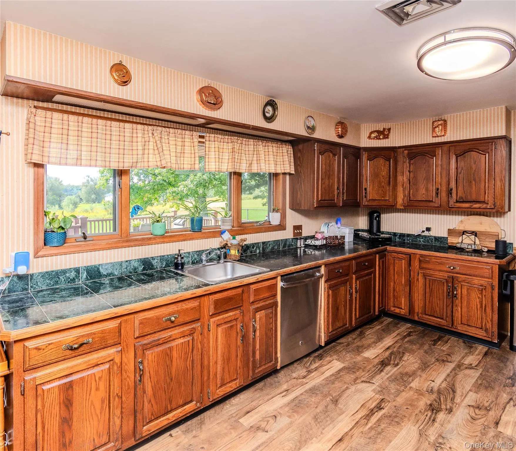 Wood-finish flooring, wood cabinetry, tile countertops, stainless steel sink, and stainless steel dishwasher Wood-finish flooring, wood cabinetry, tile countertops, stainless steel sink, and stainless steel dishwasher