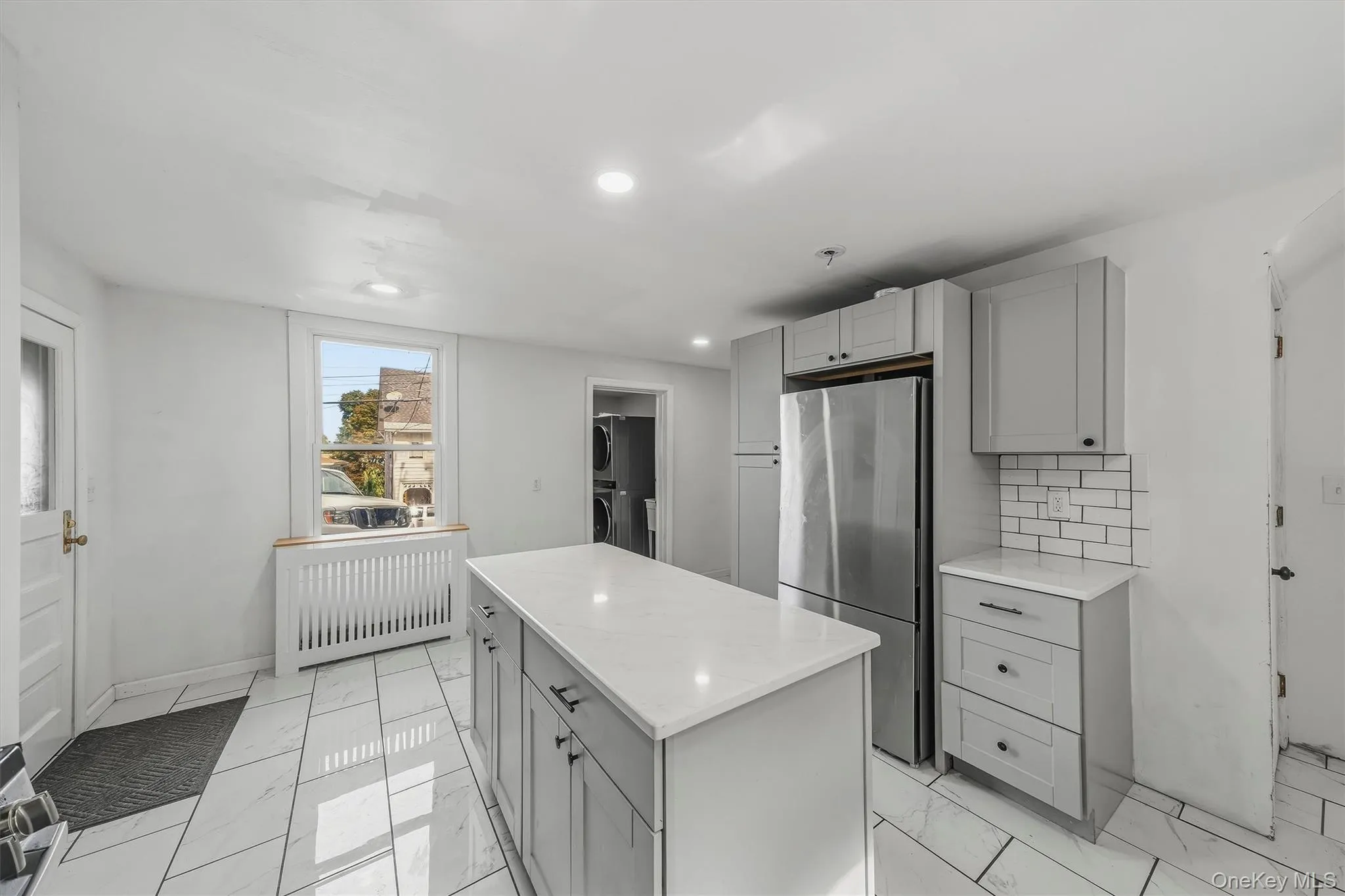 Kitchen featuring a central island with countertop, stainless steel refrigerator, light gray cabinetry, subway tile backsplash, and white tile flooring Kitchen featuring a central island with countertop, stainless steel refrigerator, light gray cabinetry, subway tile backsplash, and white tile flooring
