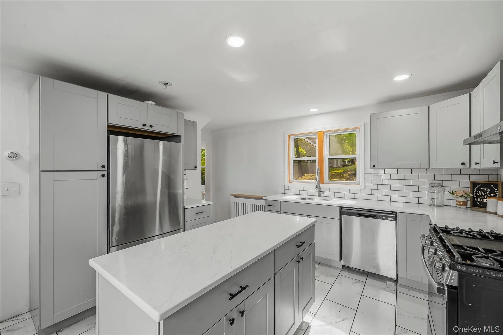 Kitchen featuring gray shaker-style cabinetry, a central island with a light-toned countertop, a subway tile backsplash, and stainless steel appliances Kitchen featuring gray shaker-style cabinetry, a central island with a light-toned countertop, a subway tile backsplash, and stainless steel appliances