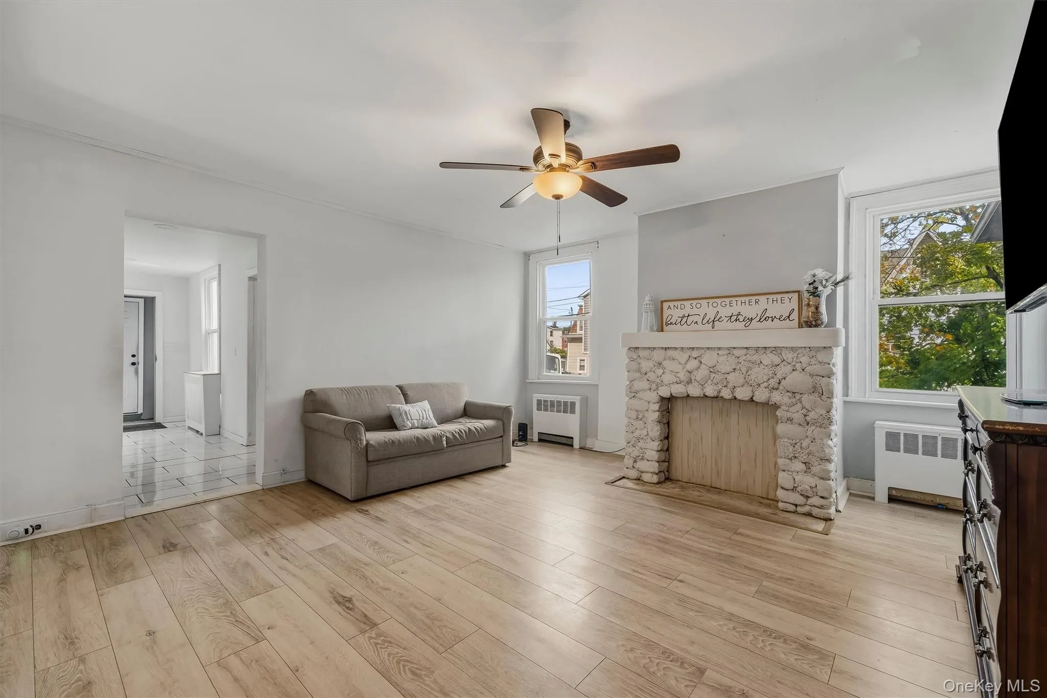 Light wood-finish flooring, stone-clad fireplace with mantel, ceiling fan with integrated lighting, and multiple windows Light wood-finish flooring, stone-clad fireplace with mantel, ceiling fan with integrated lighting, and multiple windows