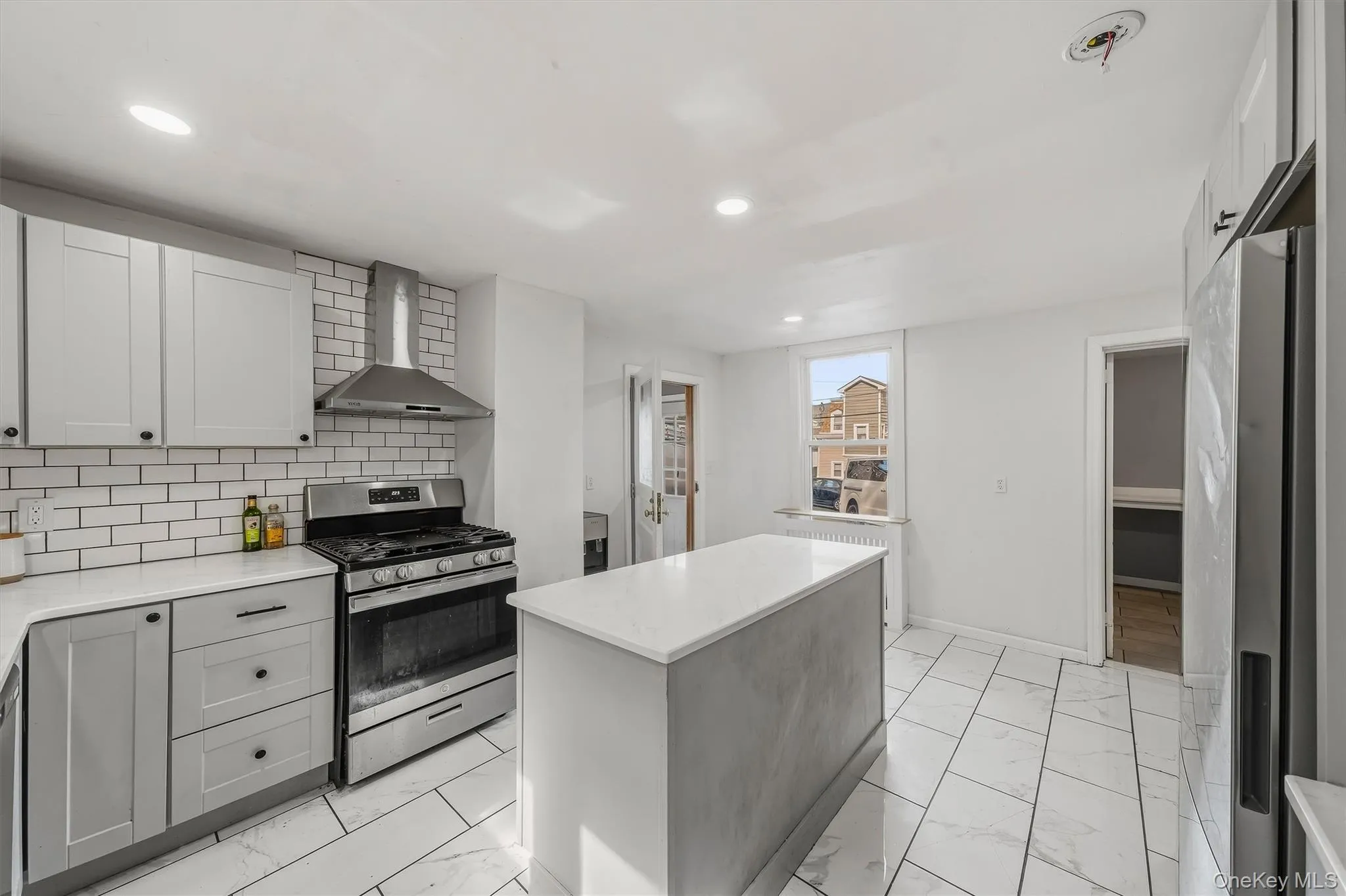 Modern kitchen featuring white subway tile backsplash, stainless steel range and hood, light-toned cabinetry, a central island, and large format floor tiling Modern kitchen featuring white subway tile backsplash, stainless steel range and hood, light-toned cabinetry, a central island, and large format floor tiling