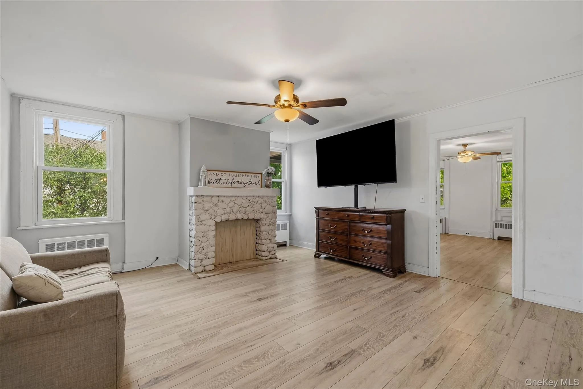 Light wood-finish flooring, stone-clad fireplace, white painted walls, and a ceiling fan with integrated lighting Light wood-finish flooring, stone-clad fireplace, white painted walls, and a ceiling fan with integrated lighting
