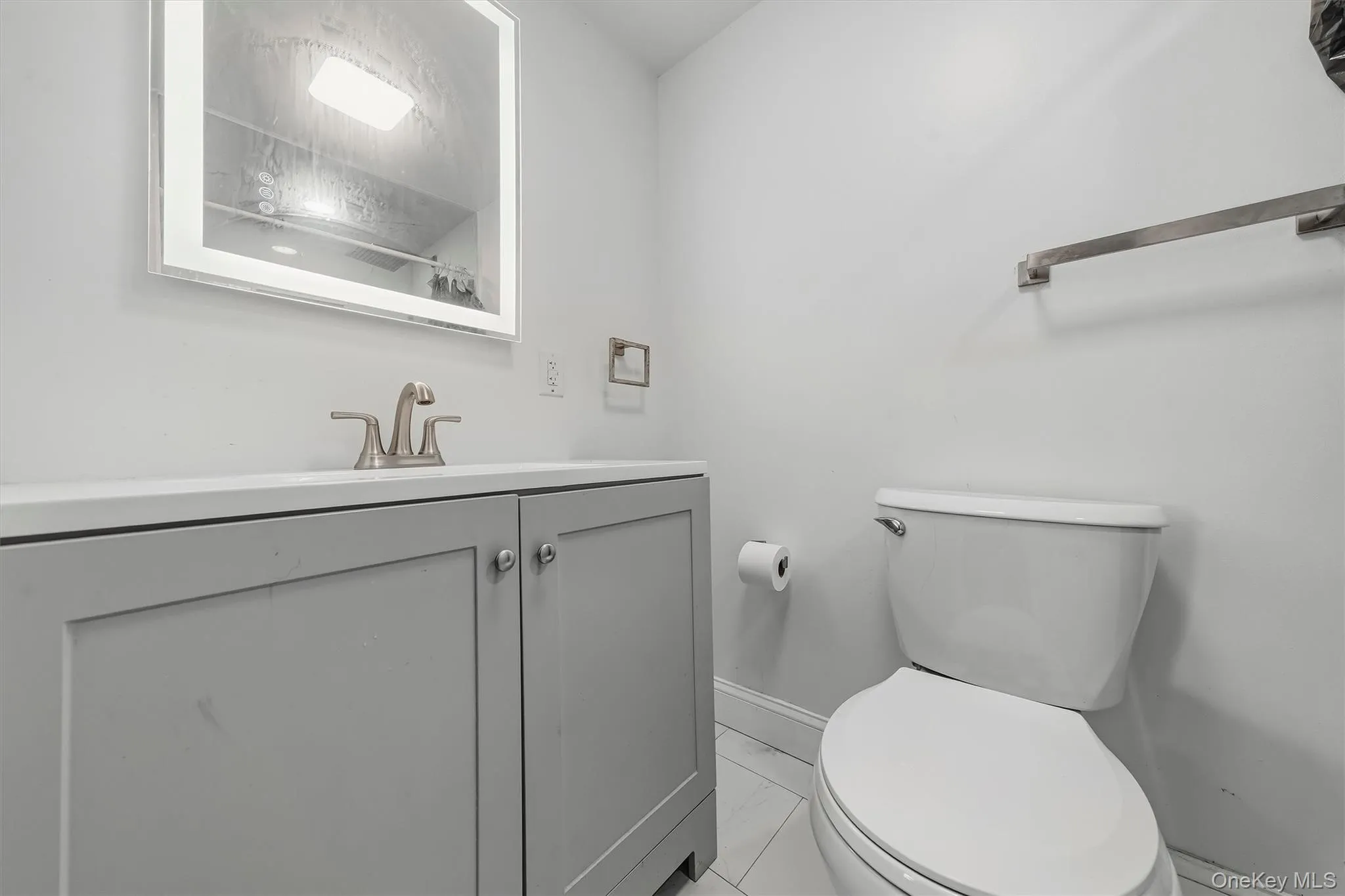 Lighted vanity mirror, gray vanity with cabinet storage, white countertop, modern brushed nickel faucet, and white tile flooring Lighted vanity mirror, gray vanity with cabinet storage, white countertop, modern brushed nickel faucet, and white tile flooring