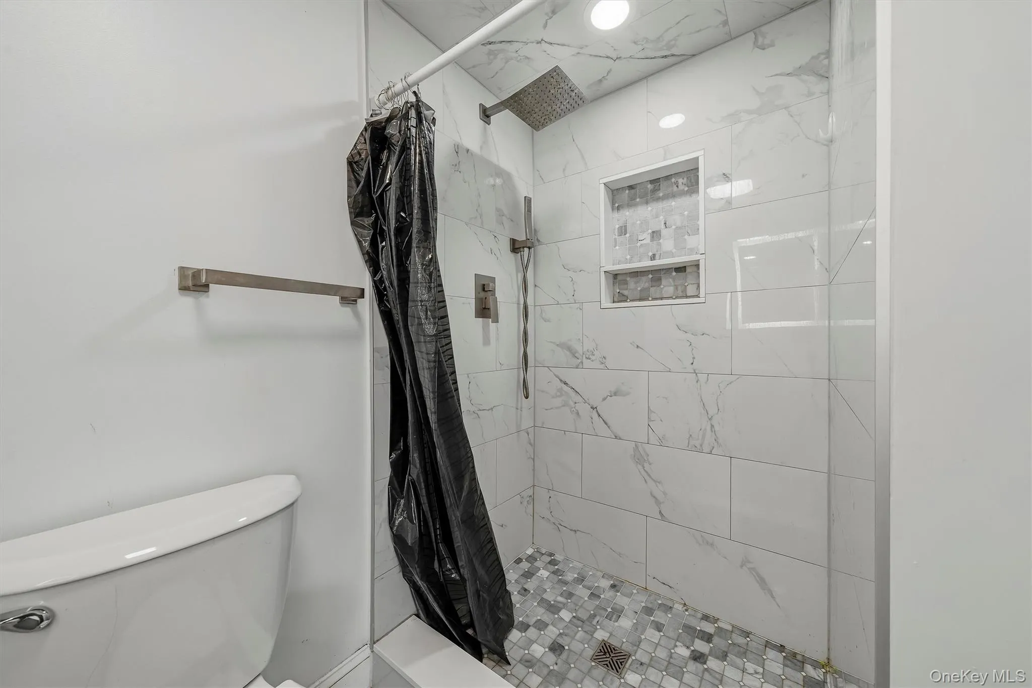 Shower featuring white marble-look subway tile, mosaic tile shower floor, built-in shower niche, and a rain shower head with an additional handheld shower fixture Shower featuring white marble-look subway tile, mosaic tile shower floor, built-in shower niche, and a rain shower head with an additional handheld shower fixture