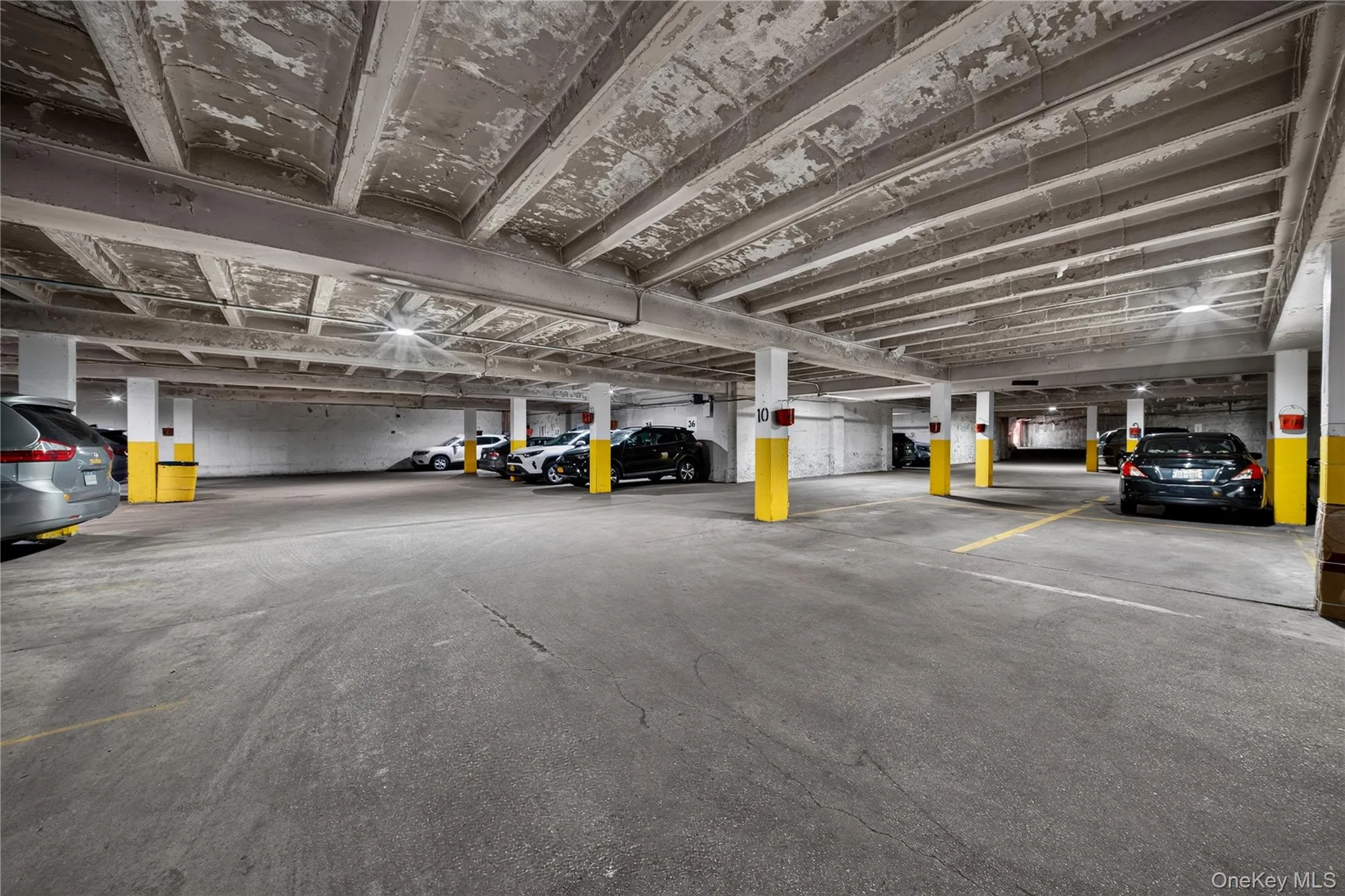 Covered parking garage featuring concrete flooring, structural columns, and overhead lighting Covered parking garage featuring concrete flooring, structural columns, and overhead lighting