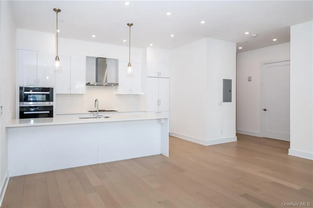 Kitchen featuring light hardwood / wood-style floors, wall chimney exhaust hood, decorative light fixtures, double oven, and white cabinets Kitchen featuring light hardwood / wood-style floors, wall chimney exhaust hood, decorative light fixtures, double oven, and white cabinets