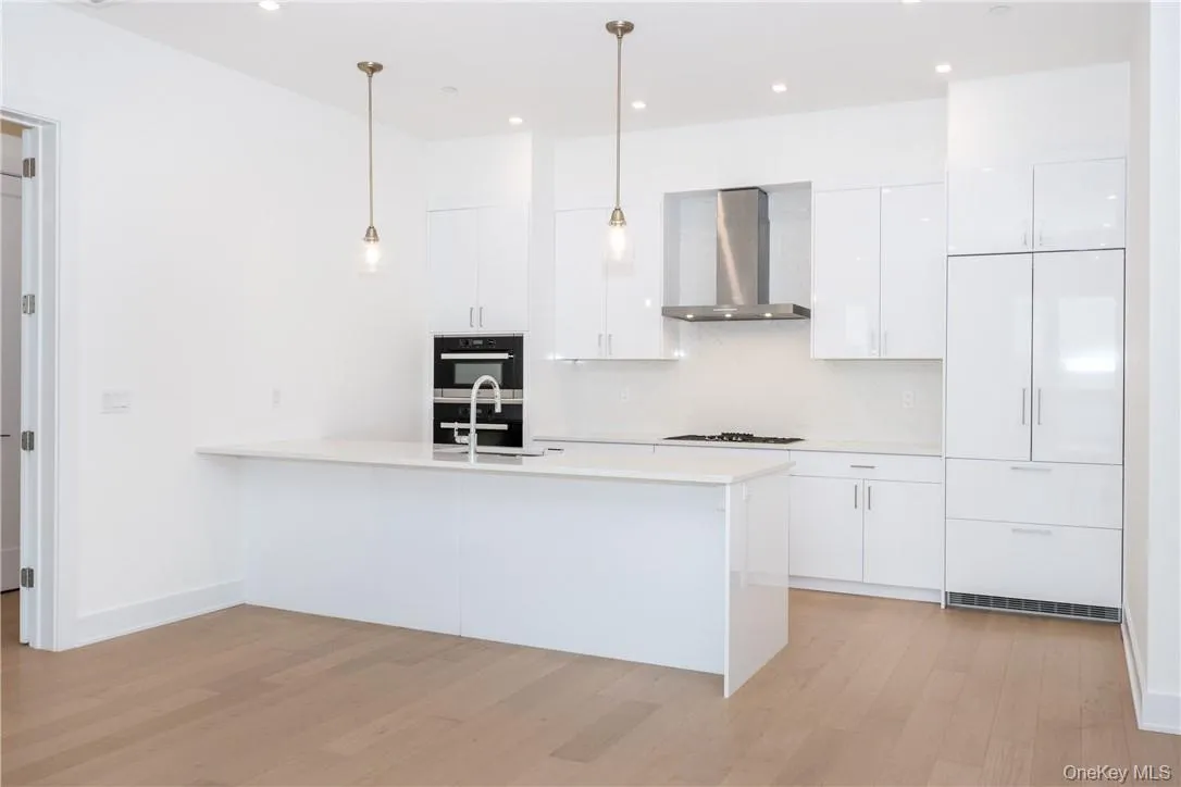 Kitchen featuring stainless steel double oven, wall chimney range hood, white cabinetry, light wood-type flooring, and decorative light fixtures Kitchen featuring stainless steel double oven, wall chimney range hood, white cabinetry, light wood-type flooring, and decorative light fixtures