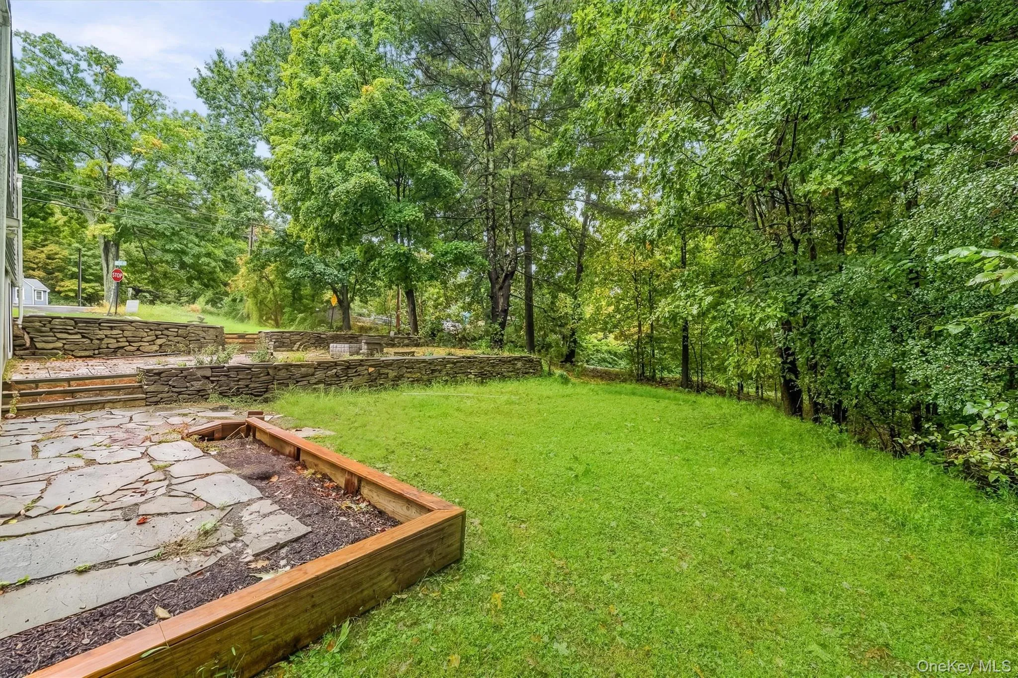 Expansive green lawn surrounded by mature trees, featuring a flagstone patio with a raised wood-finish planter box Expansive green lawn surrounded by mature trees, featuring a flagstone patio with a raised wood-finish planter box