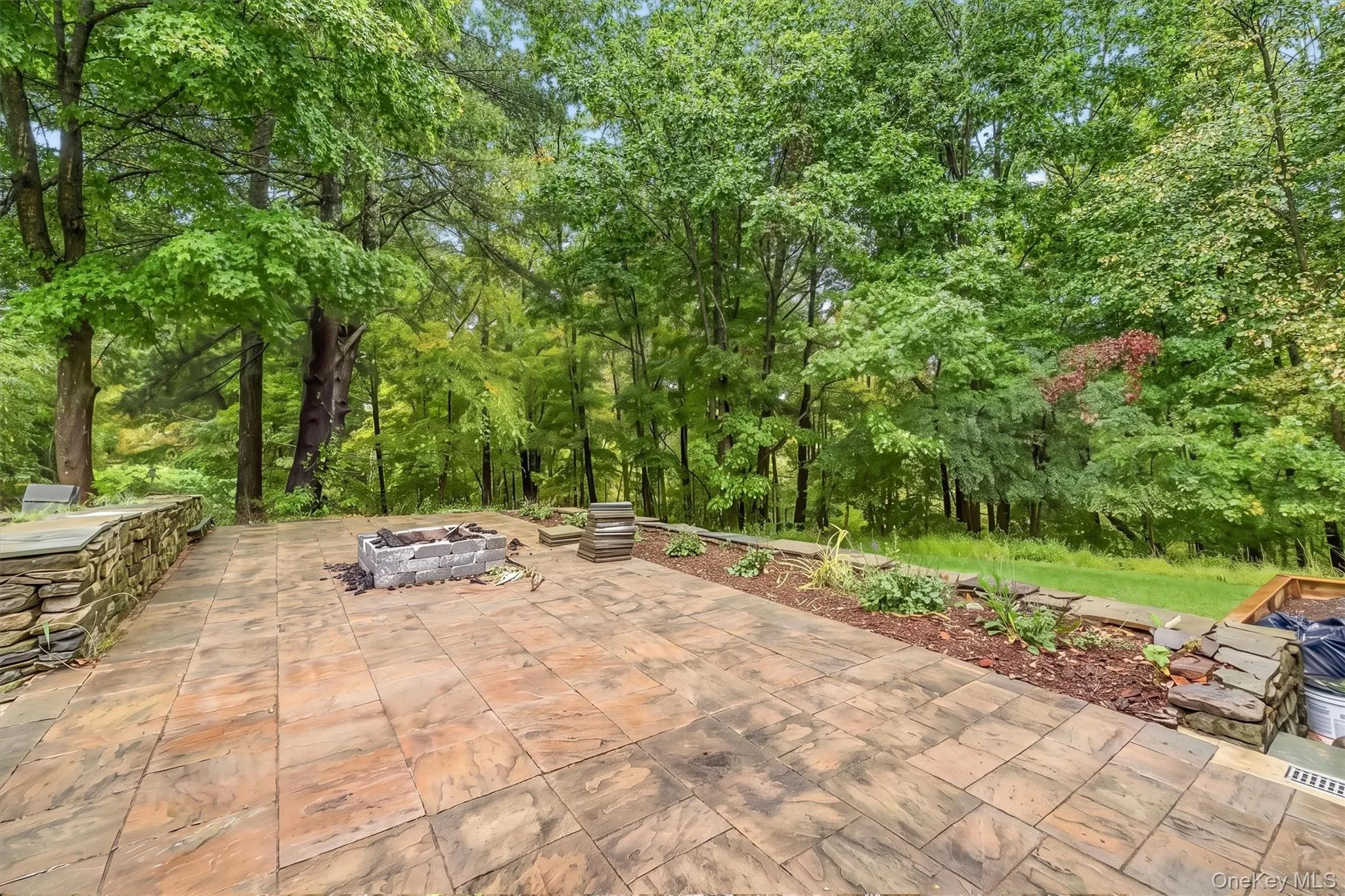 Expansive paver patio with a stone fire pit, surrounded by mature trees, stone retaining walls, and planted garden beds Expansive paver patio with a stone fire pit, surrounded by mature trees, stone retaining walls, and planted garden beds