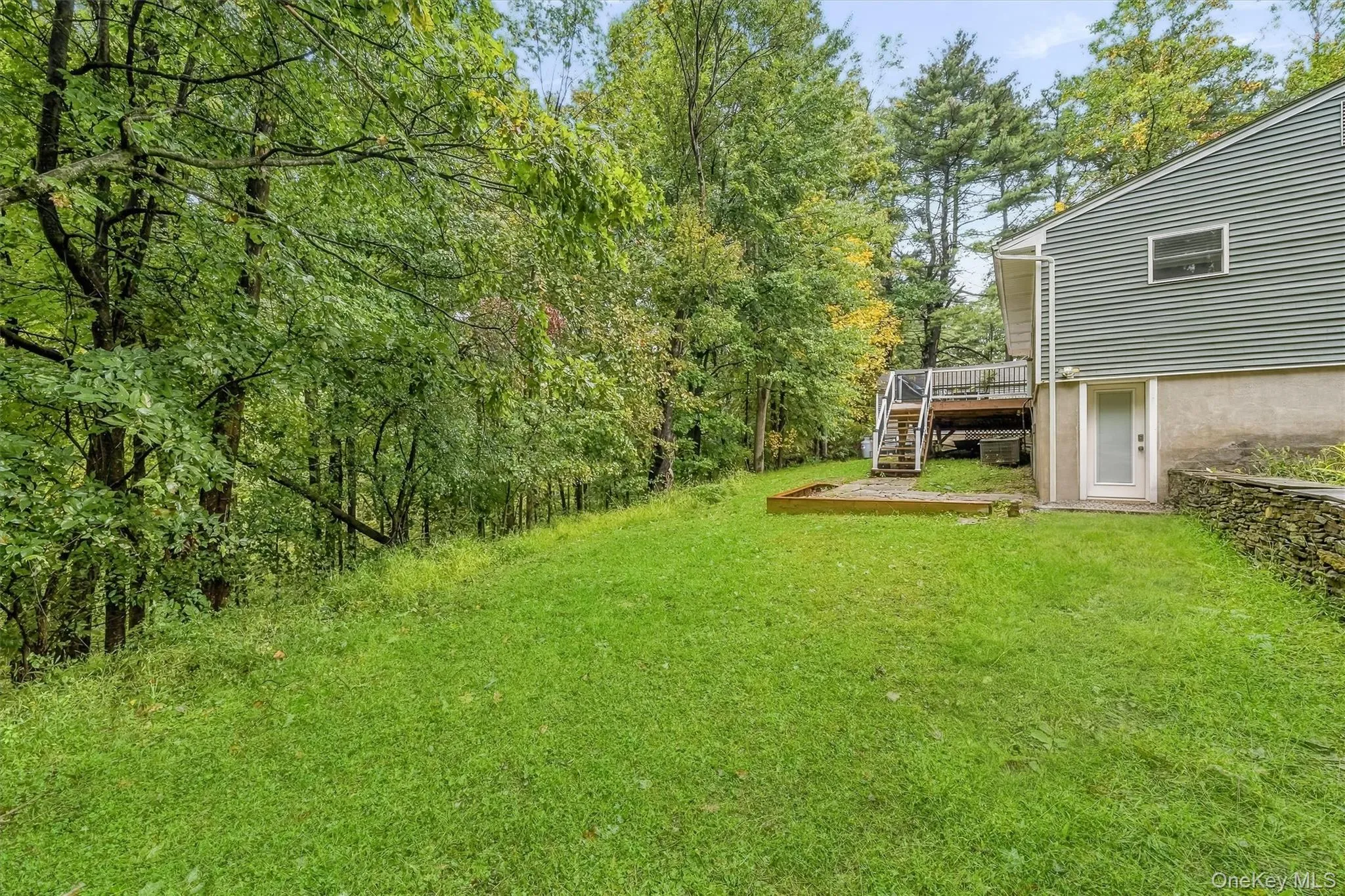 Expansive green lawn, mature trees, multi-level exterior deck, natural stone retaining wall, and a ground-level exterior door Expansive green lawn, mature trees, multi-level exterior deck, natural stone retaining wall, and a ground-level exterior door
