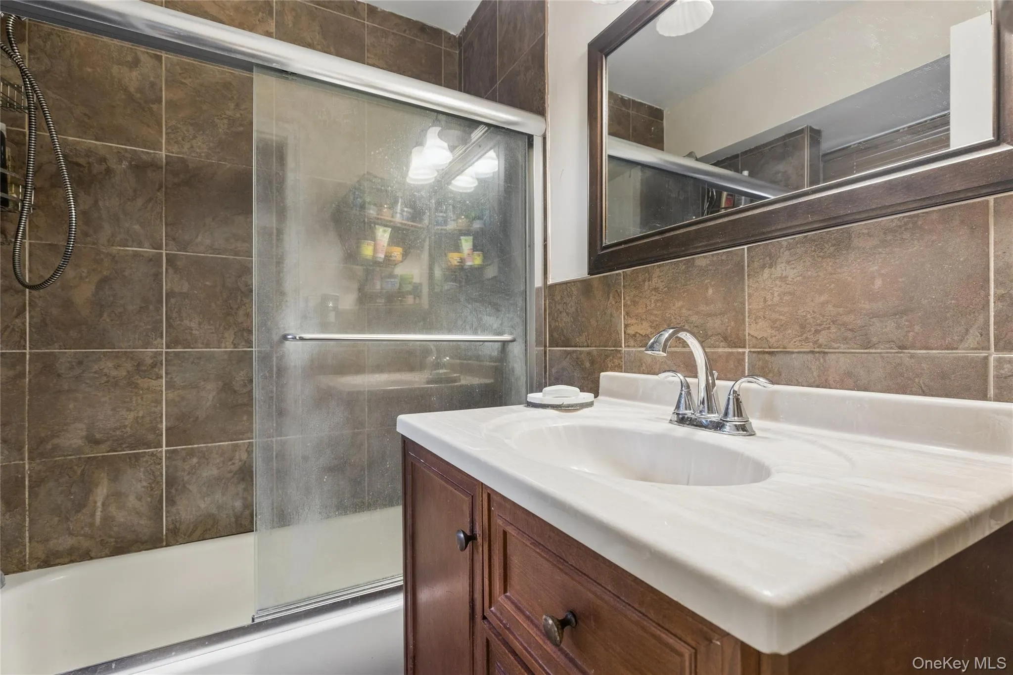 Full-height wall tiling, tub with sliding glass shower door, integrated sink with chrome faucet, wood-finish vanity, and framed mirror Full-height wall tiling, tub with sliding glass shower door, integrated sink with chrome faucet, wood-finish vanity, and framed mirror