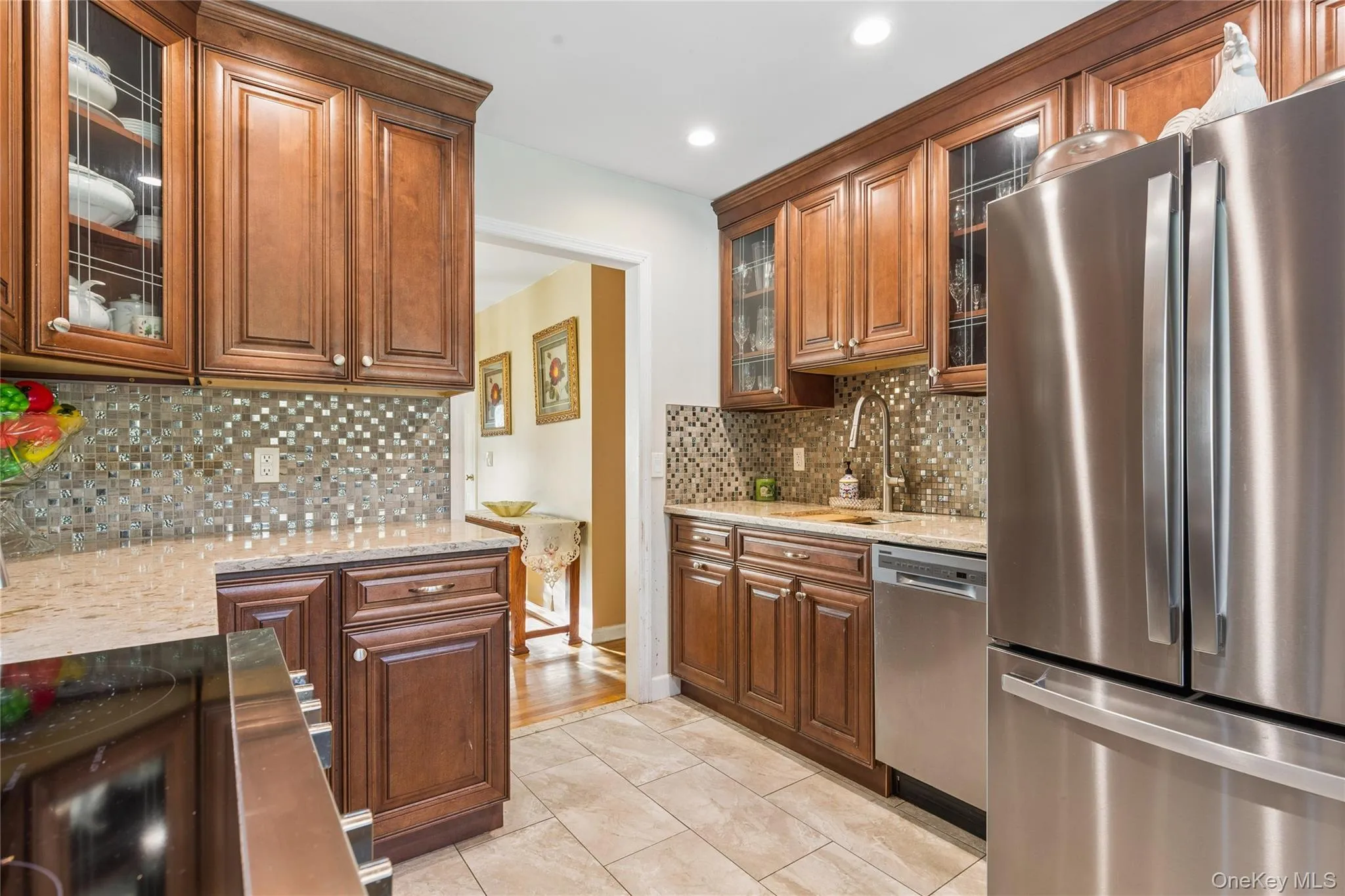 Elegant galley kitchen featuring rich wood cabinetry with glass-front upper storage, stainless steel French-door refrigerator and dishwasher, mosaic tile backsplash, and neutral ceramic tile flooring throughout Elegant galley kitchen featuring rich wood cabinetry with glass-front upper storage, stainless steel French-door refrigerator and dishwasher, mosaic tile backsplash, and neutral ceramic tile flooring throughout