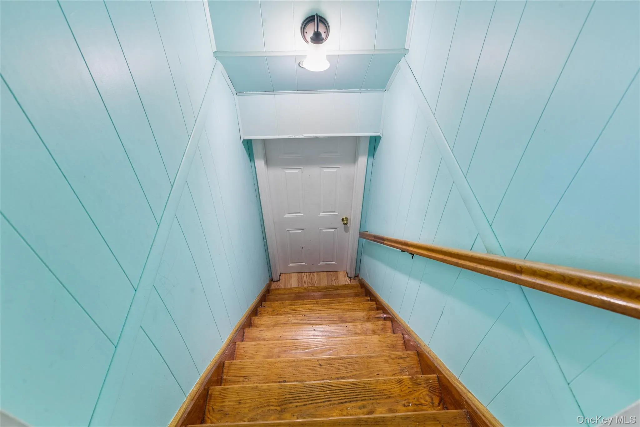 Wood plank stairs with a wood handrail, paneling wall treatment, a panel door, and a flush mount light fixture Wood plank stairs with a wood handrail, paneling wall treatment, a panel door, and a flush mount light fixture