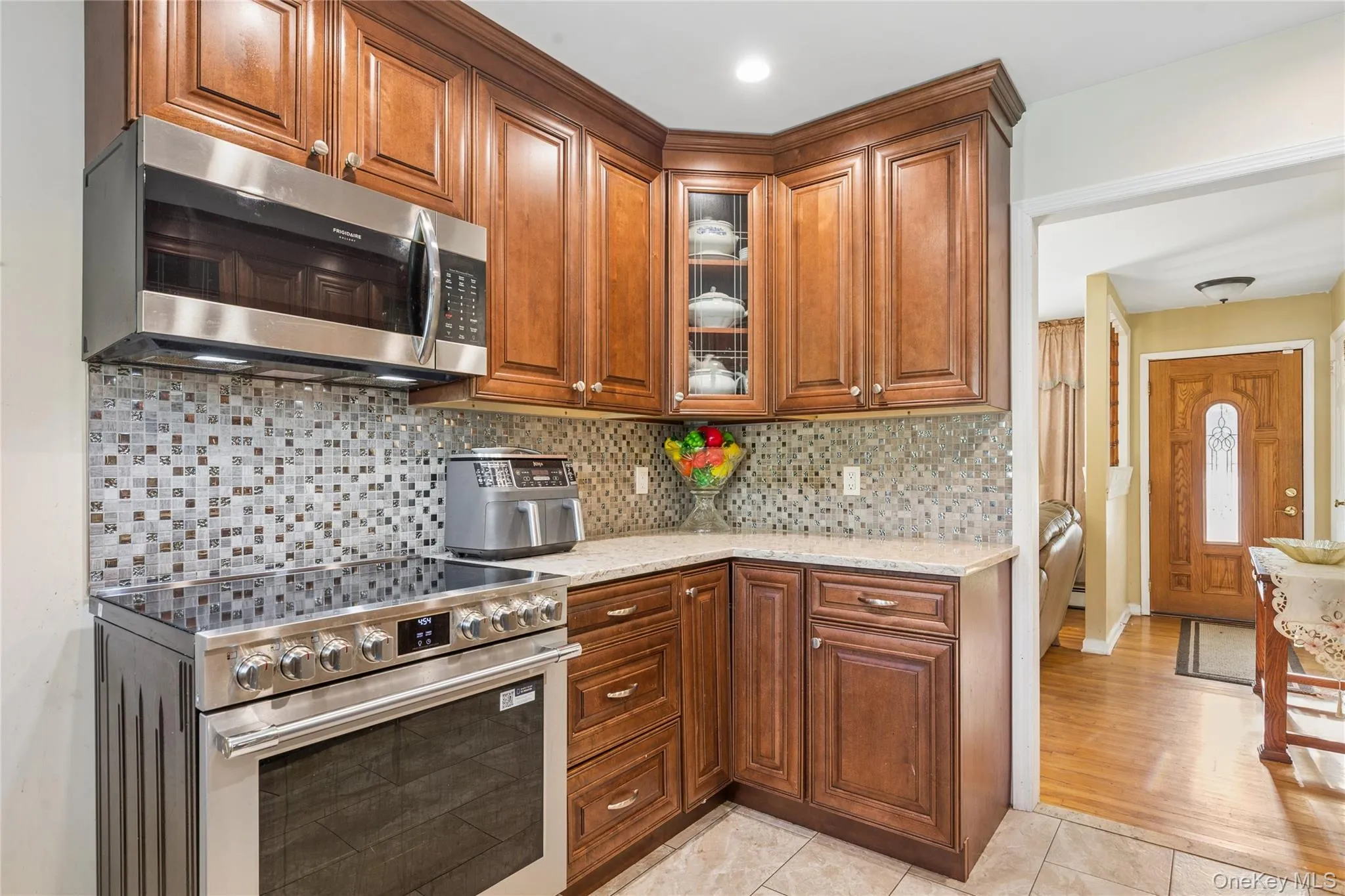 Rich wood cabinetry with a corner glass-front display, stainless steel appliances, mosaic tile backsplash, and light-toned countertop Rich wood cabinetry with a corner glass-front display, stainless steel appliances, mosaic tile backsplash, and light-toned countertop