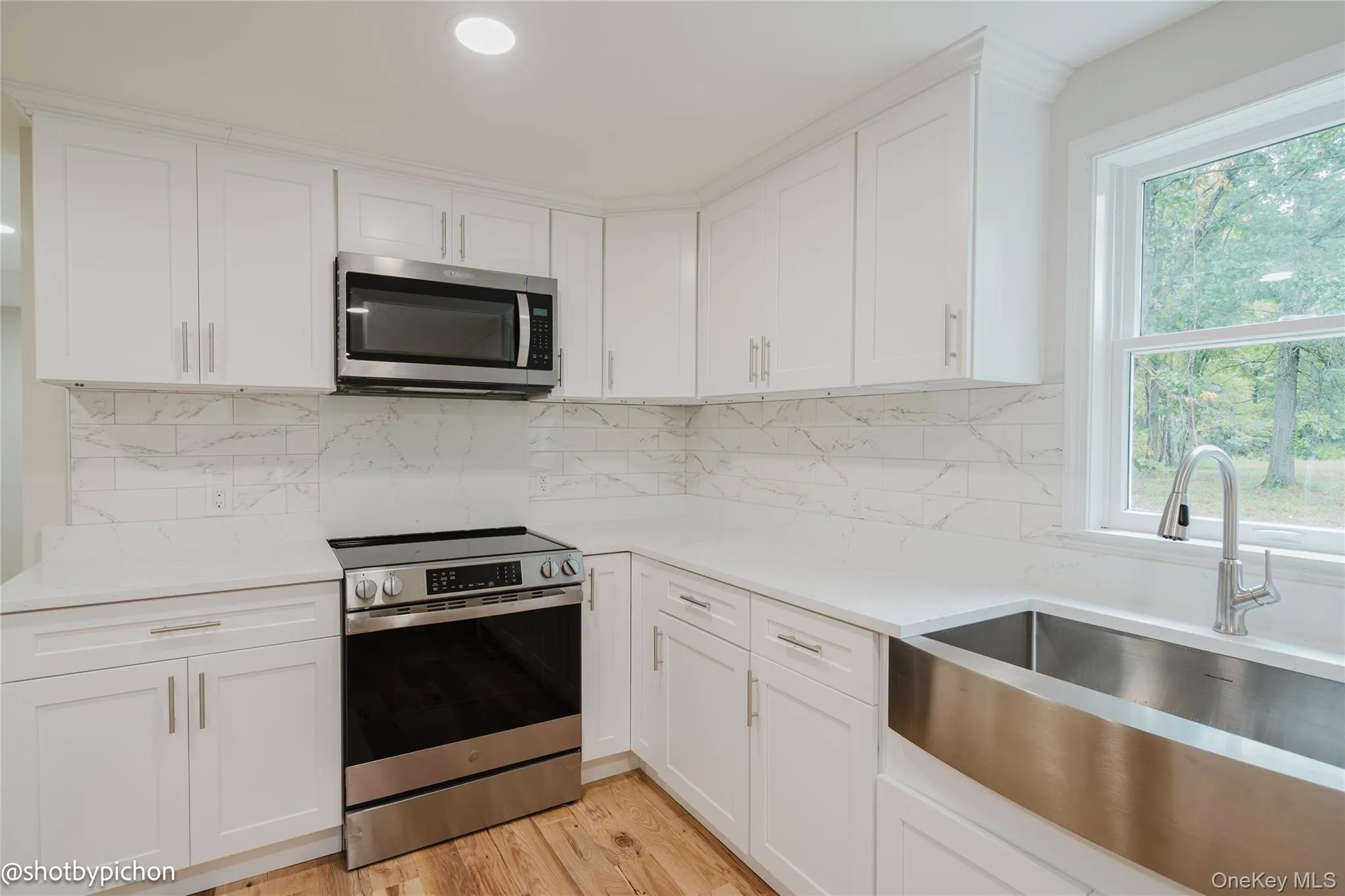 White shaker-style cabinetry with stainless steel hardware, white countertops, marble-patterned subway tile backsplash, stainless steel range, and stainless steel farmhouse sink White shaker-style cabinetry with stainless steel hardware, white countertops, marble-patterned subway tile backsplash, stainless steel range, and stainless steel farmhouse sink